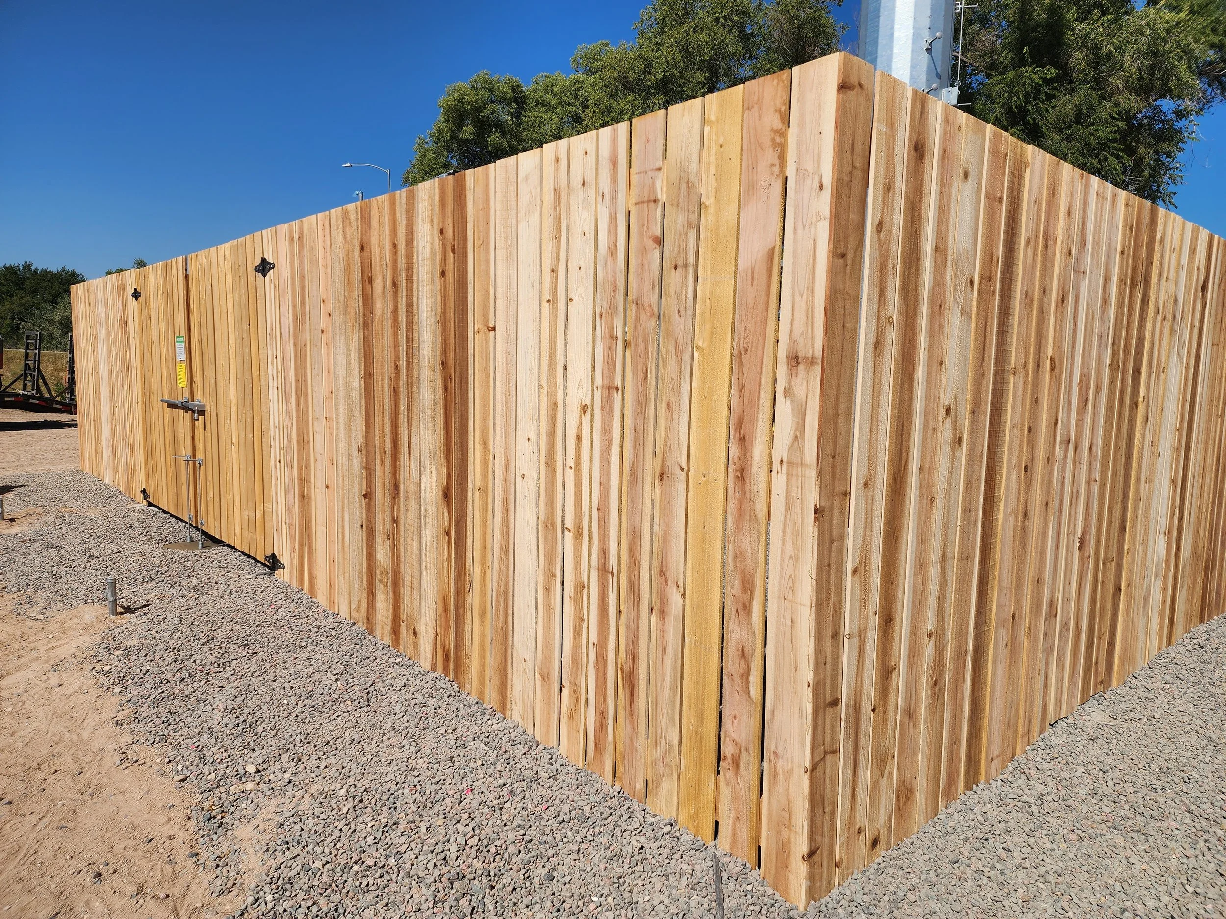 A tall wooden fence with vertical planks, set against a blue sky and green trees in the background, with a gravel surface in front.