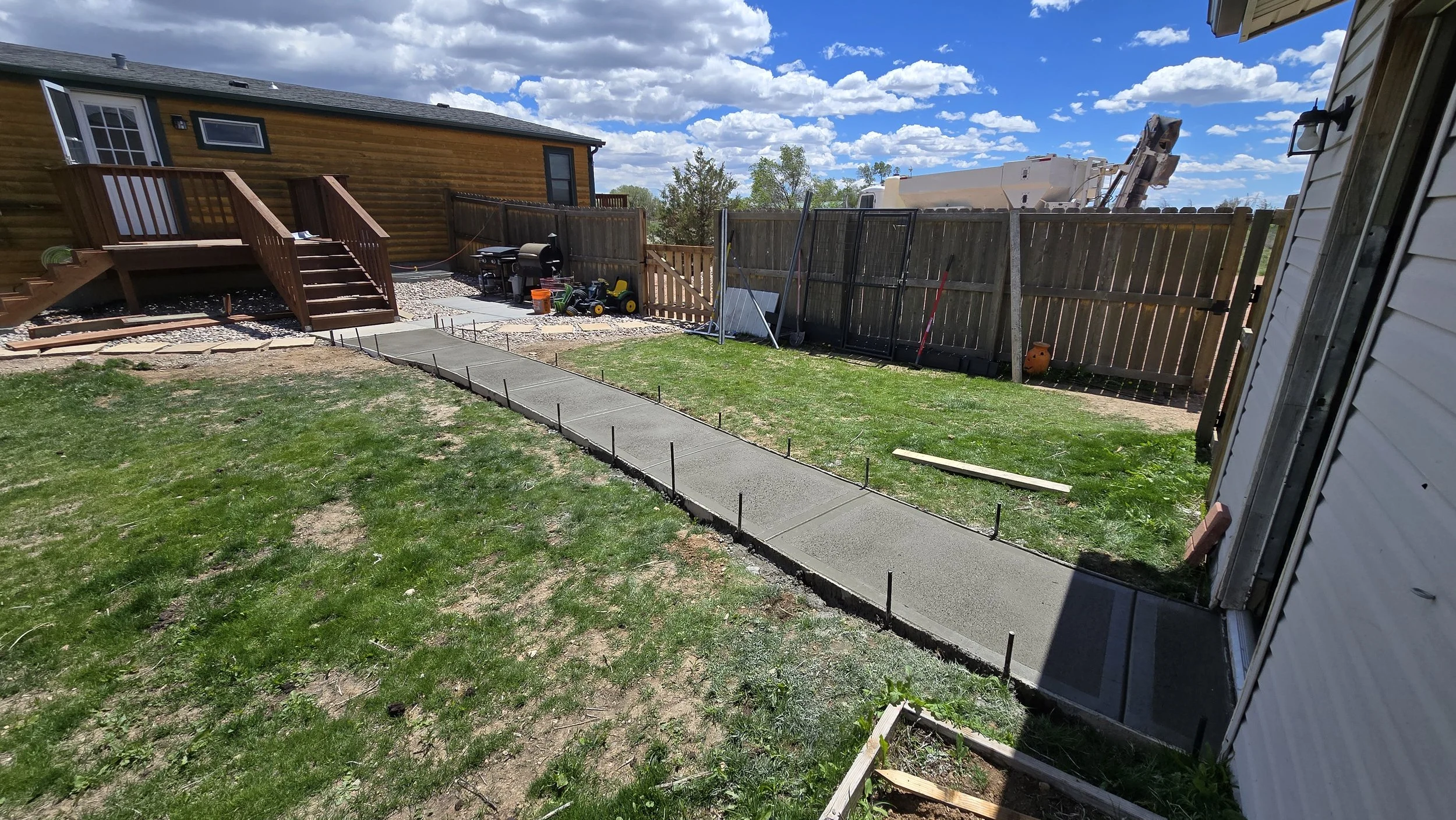 A backyard with a concrete sidewalk being newly poured, construction stakes along the edges, a grassy lawn, a wooden fence, a deck with stairs, and a cloudy sky.