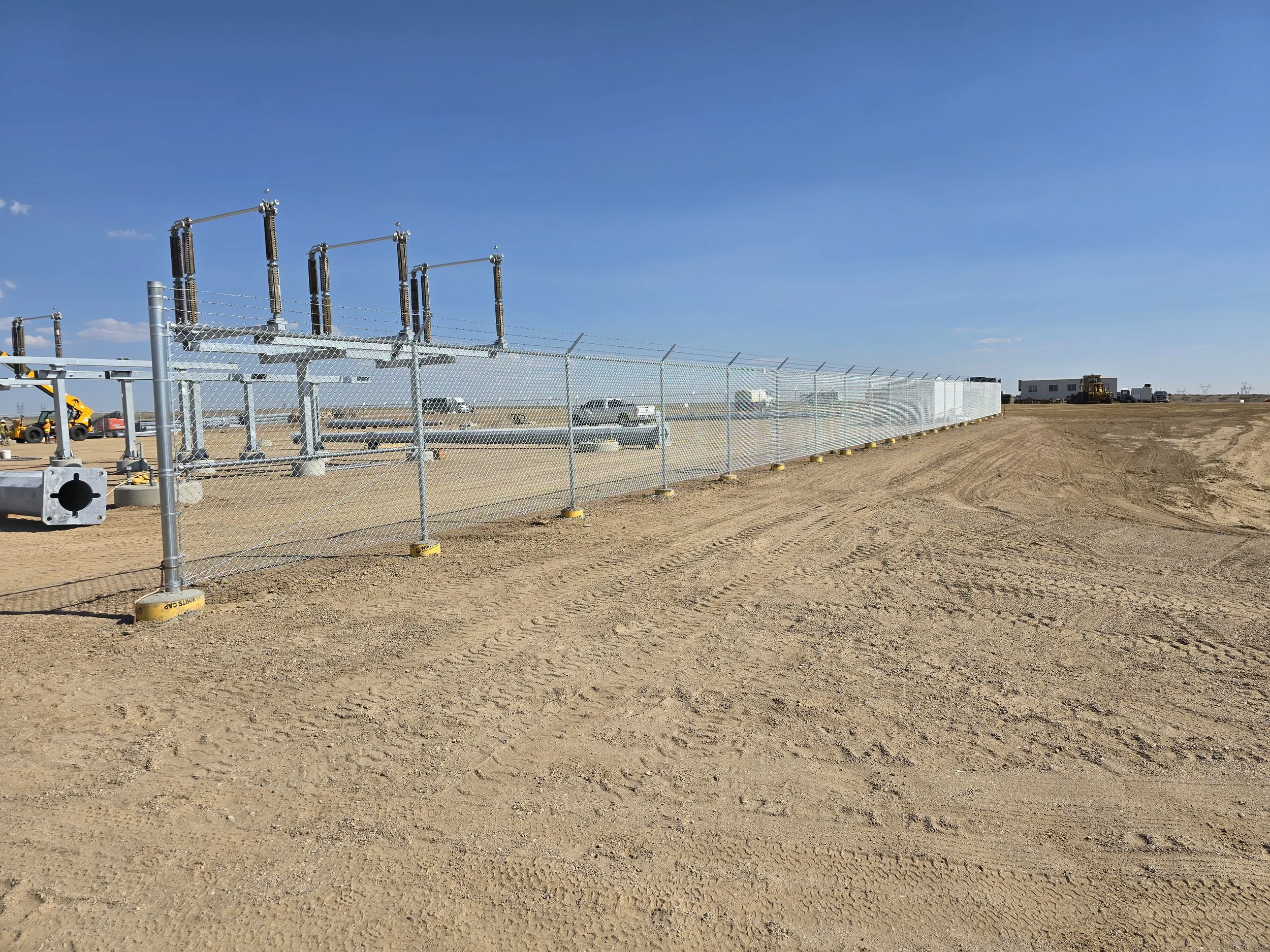 A fenced-off area with electrical infrastructure and vehicles on a flat, sandy landscape under a clear blue sky.