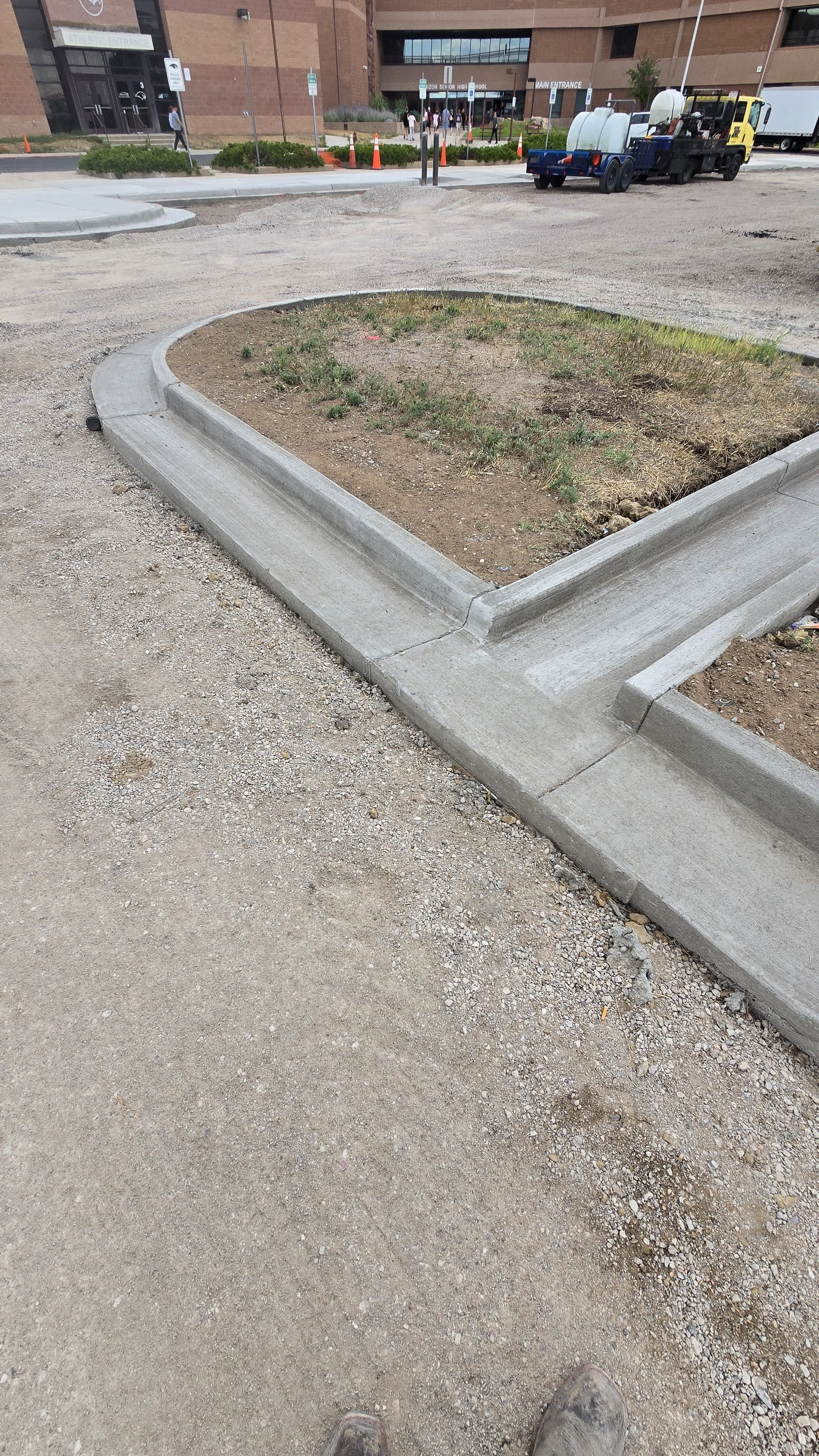 Concrete curbed planting area with grass and dirt, part of a parking lot at a hospital or medical center entrance, with a large building and people in the background.