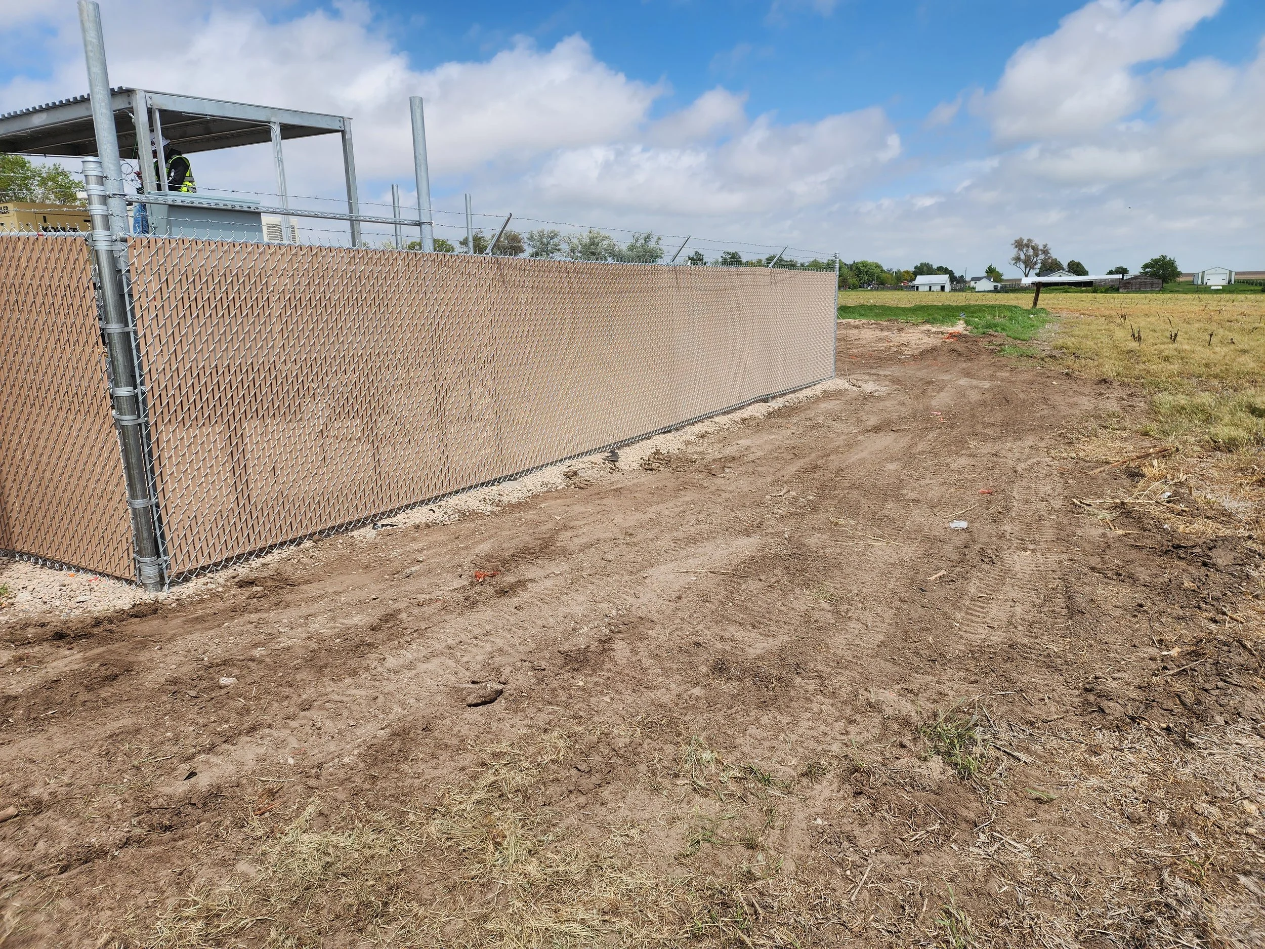 A chain-link fence with privacy slats along a dirt path, with structures and open fields in the background under a partly cloudy sky.