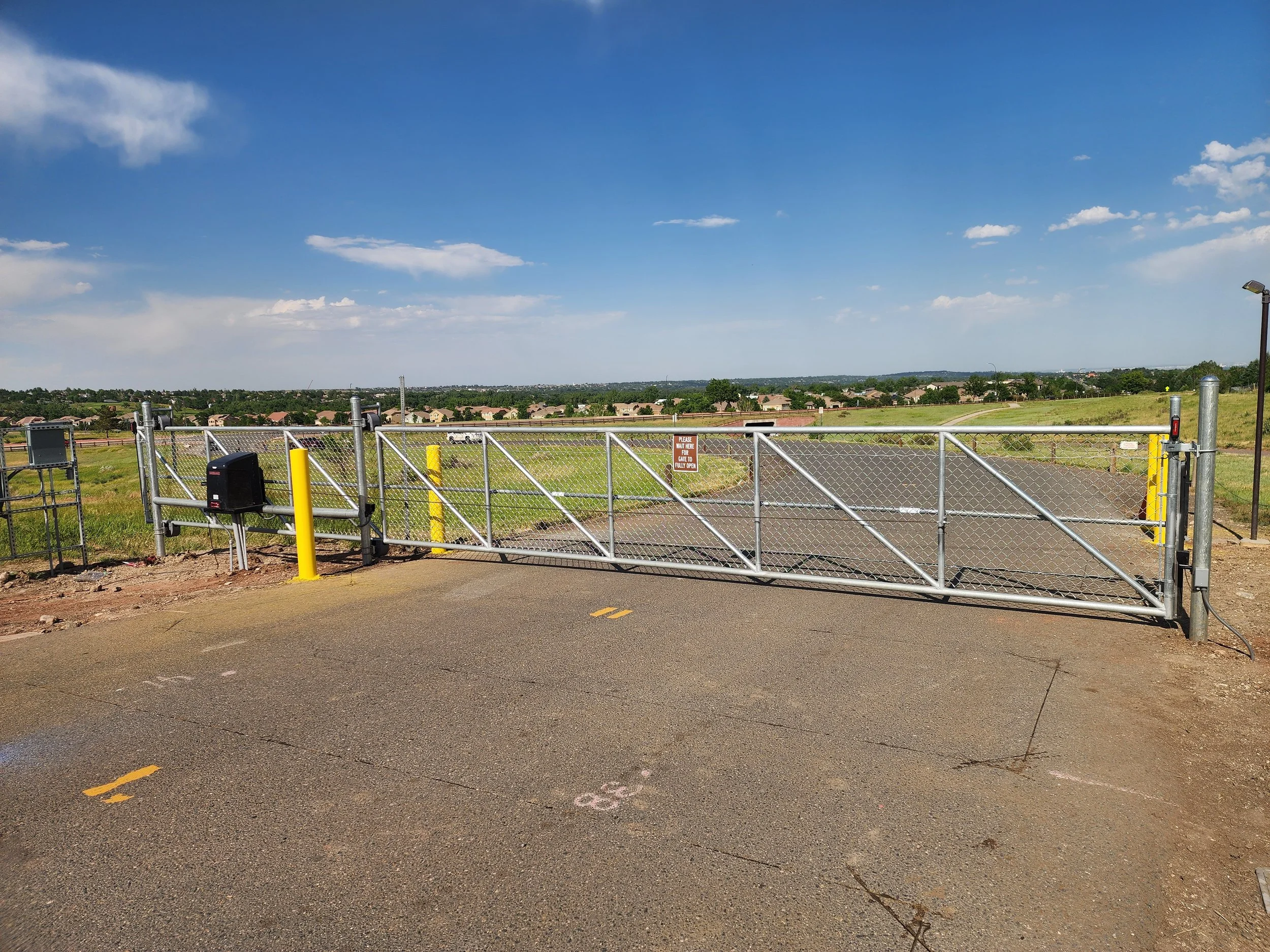 A closed metal gate with a lock, yellow posts, and a sign that says 'Please keep gate closed.' surrounding a paved area with a rural landscape and houses in the background under a blue sky with scattered clouds.
