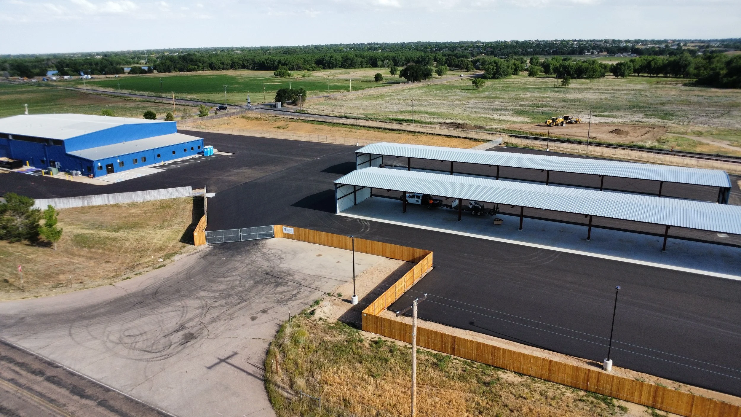 Aerial view of a new industrial or commercial building with parking area, covered storage area, and surrounding fields with construction in progress.