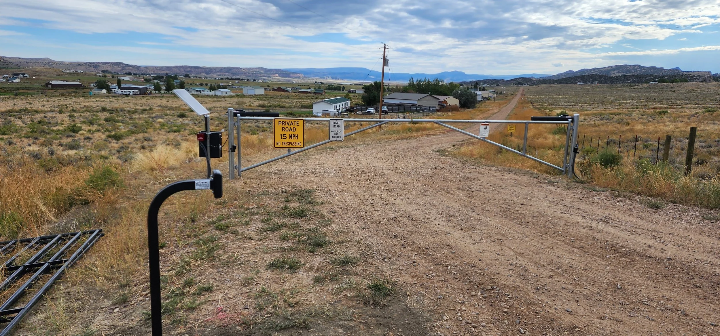 A rural dirt road blocked by a metal gate with signs indicating private property, no trespassing, and a speed limit of 15 mph. In the background, there are houses and mountains under a partly cloudy sky.