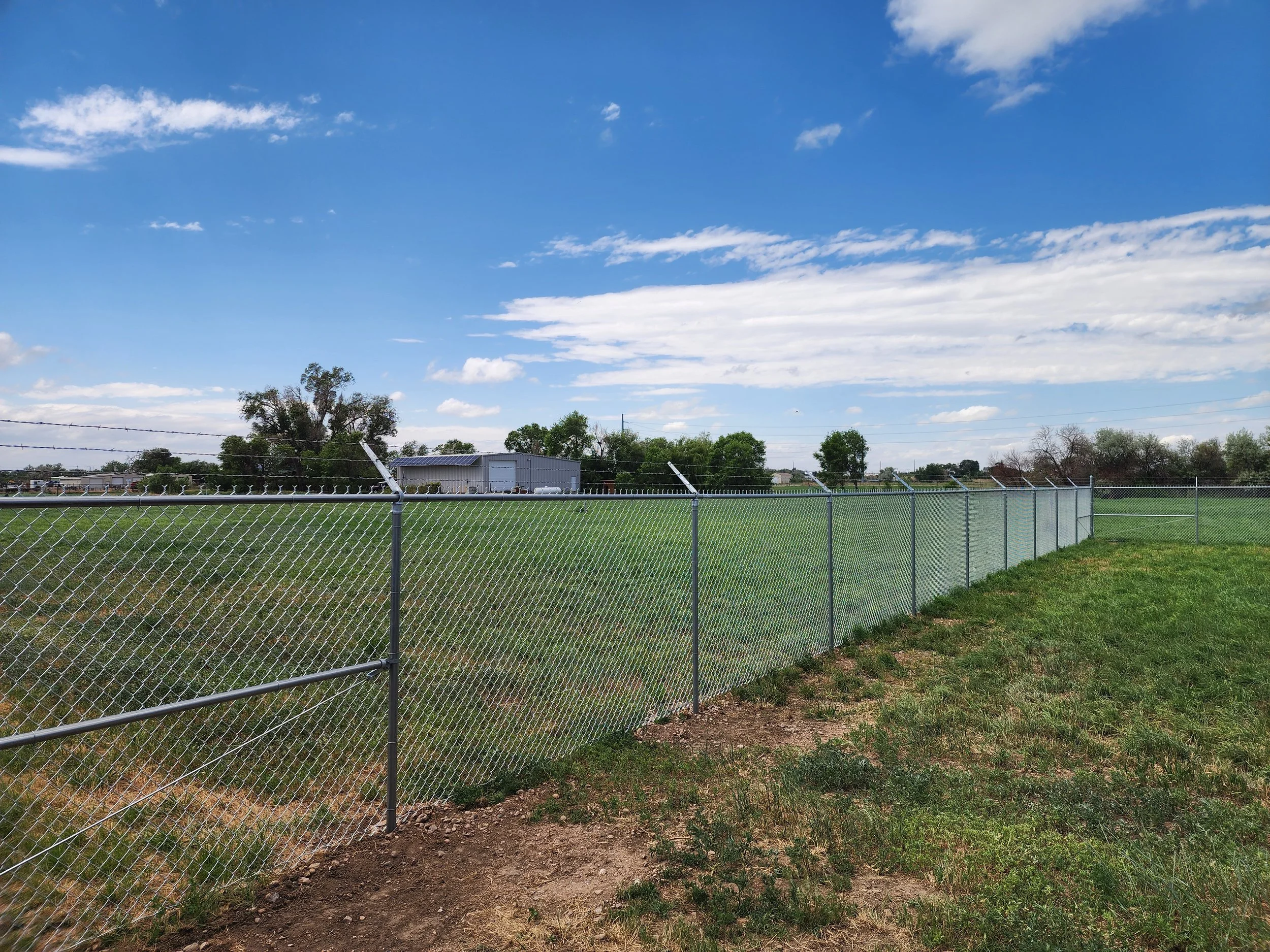 A chain-link fence running beside a grassy field under a blue sky with scattered clouds.