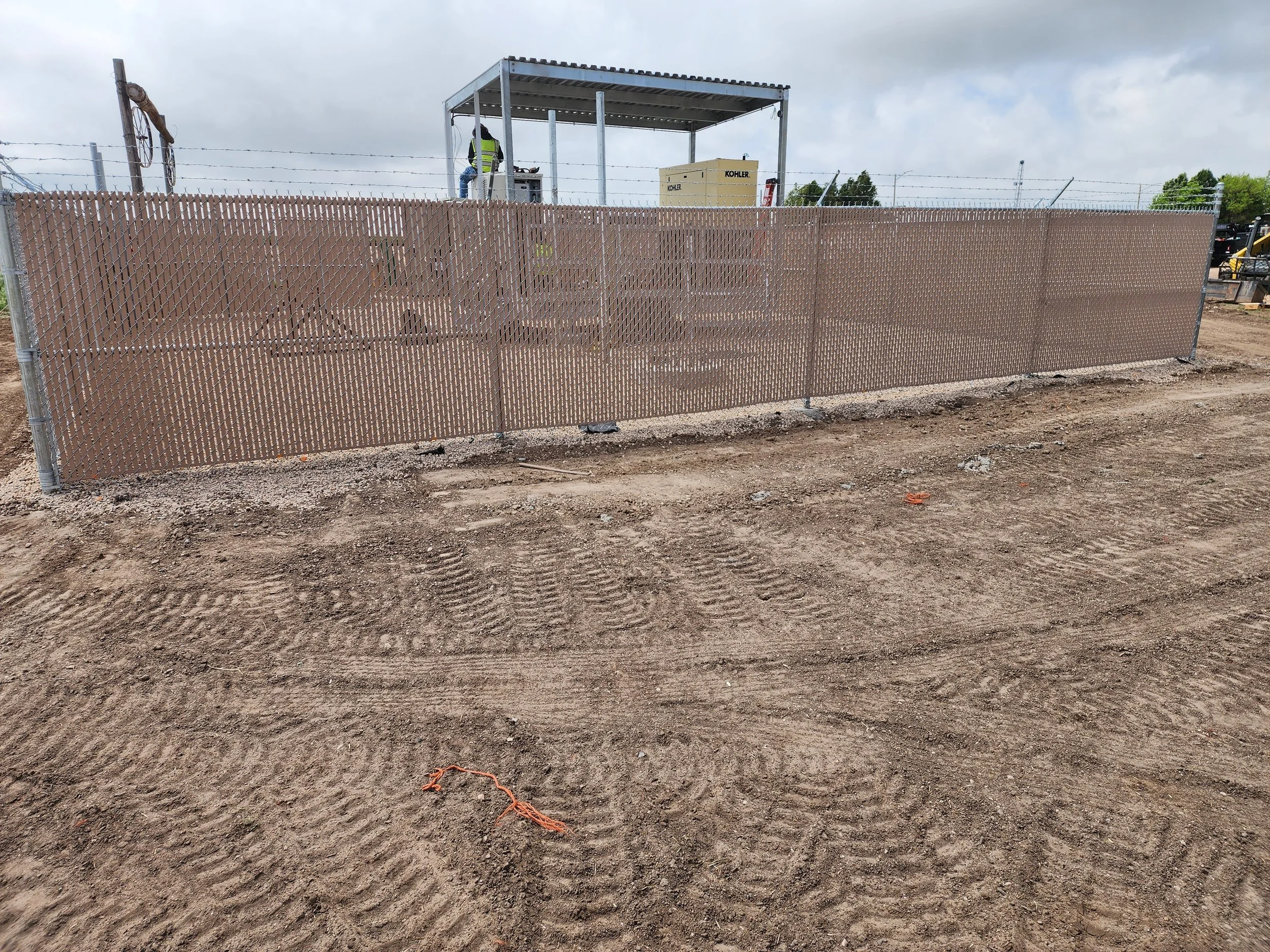 Construction site with a chain-link fence, dirt ground with tire marks, and a small structure with a worker in a yellow safety vest.