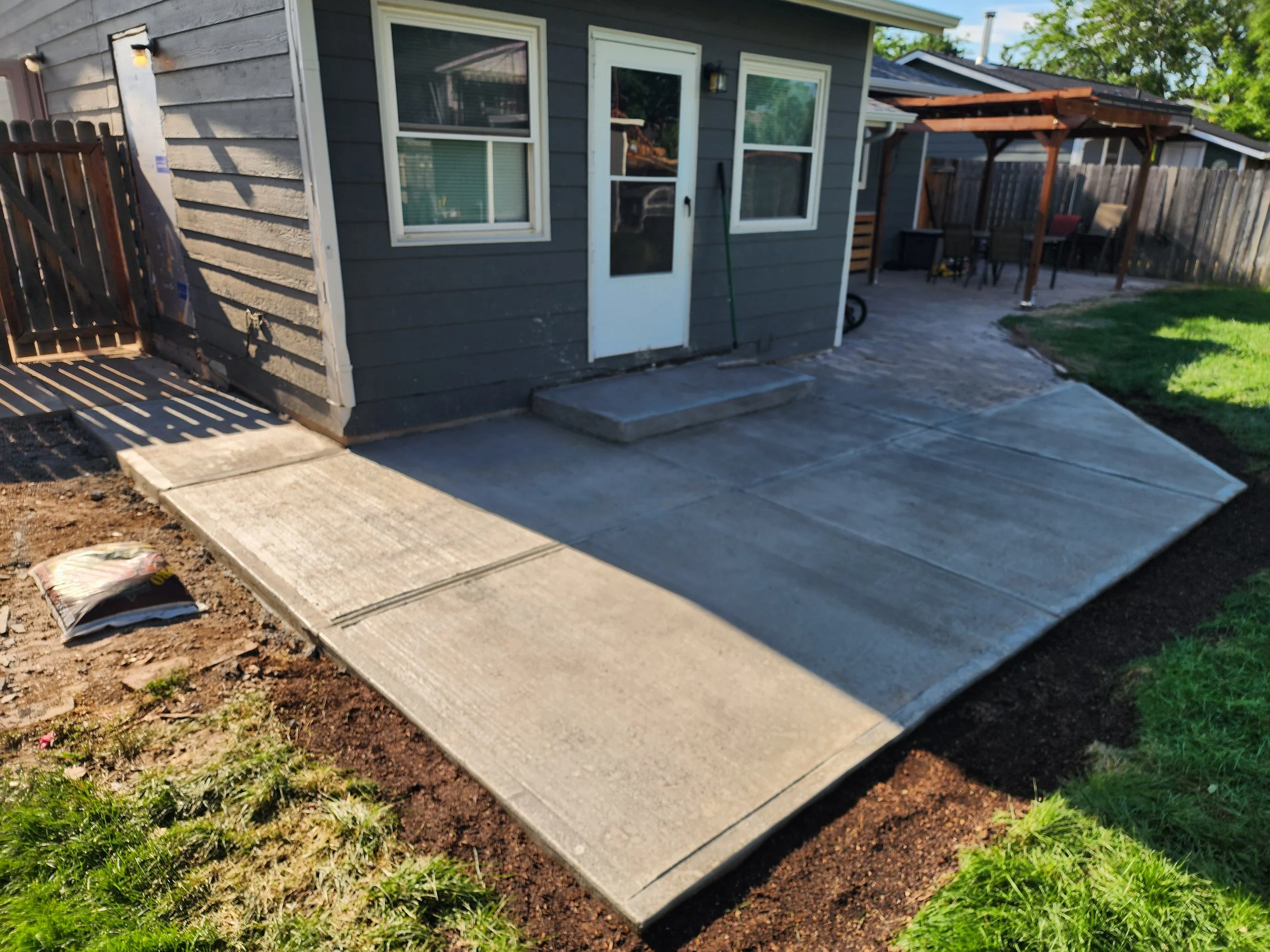Newly poured concrete patio and sidewalk in a backyard, with a house featuring gray siding, white trim, and a glass door, bordered by grass and a wooden fence.