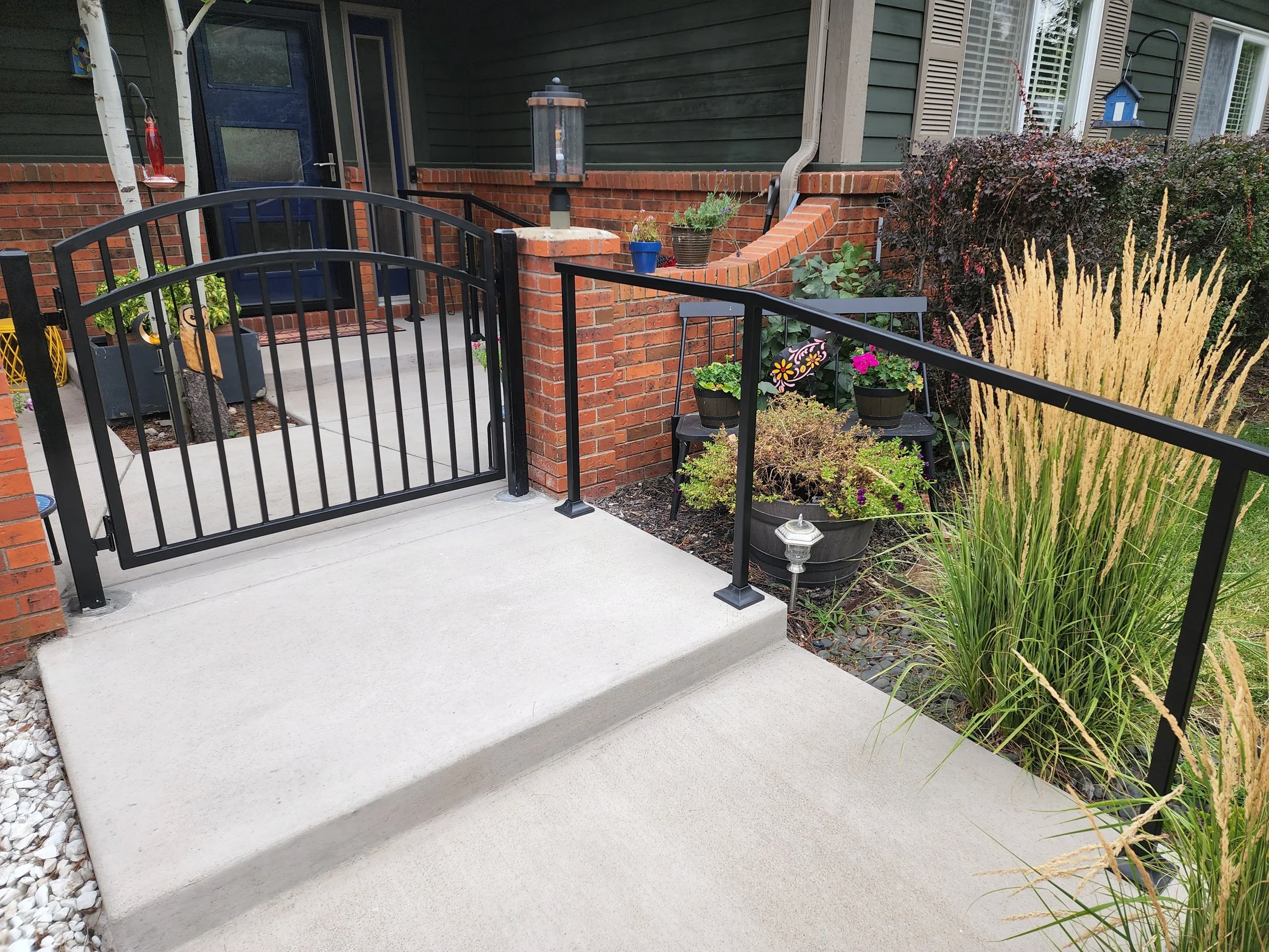 Front porch with concrete steps, black metal railing, potted plants, and landscaping with ornamental grasses and shrubs.