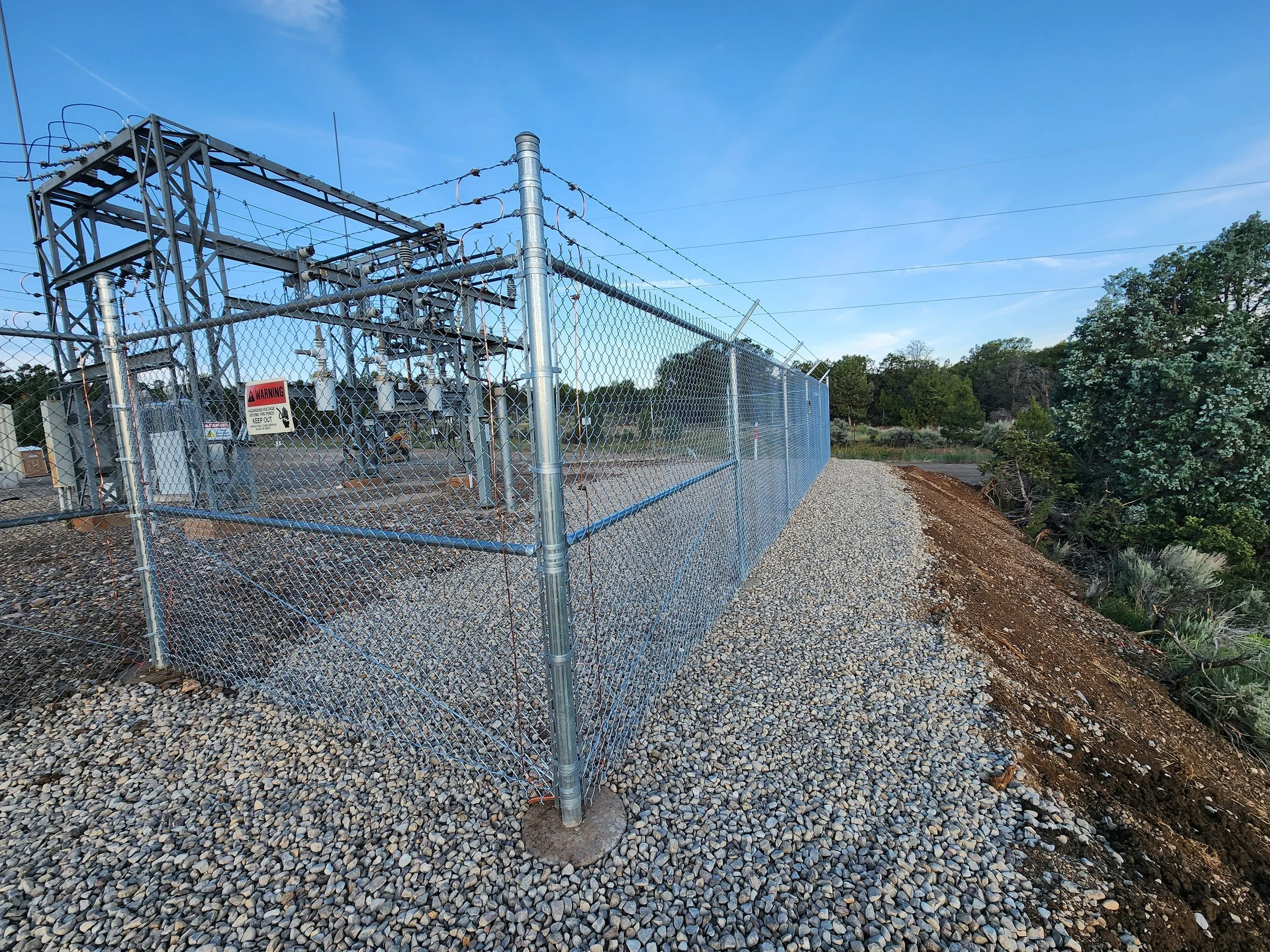 A secured electrical substation surrounded by a chain-link fence with warning signs, set in a rural area with trees and blue sky in the background.