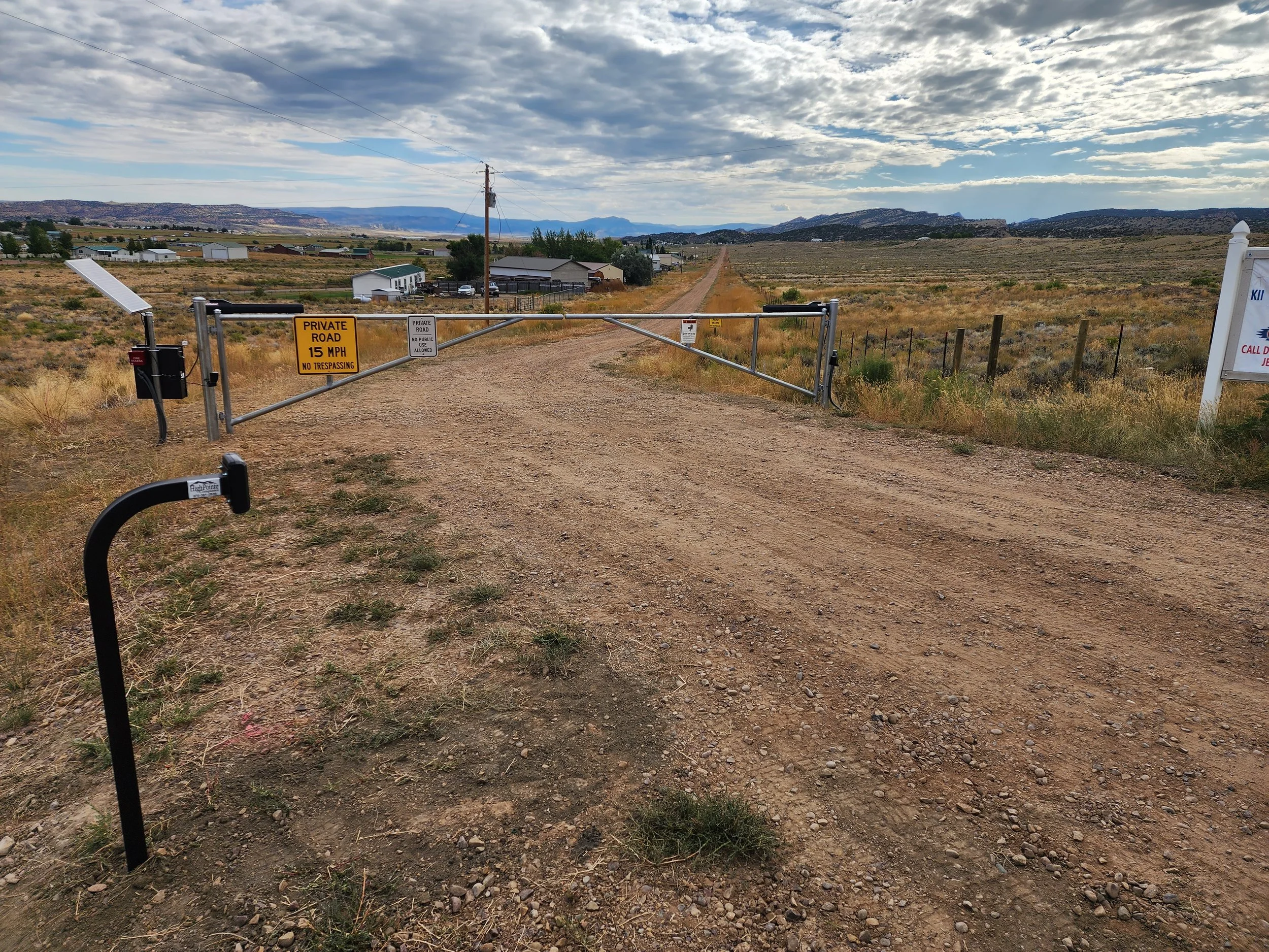 Dirt road blocked by a metal gate with signs, in a rural area with fields and mountains in the distance under a cloudy sky.