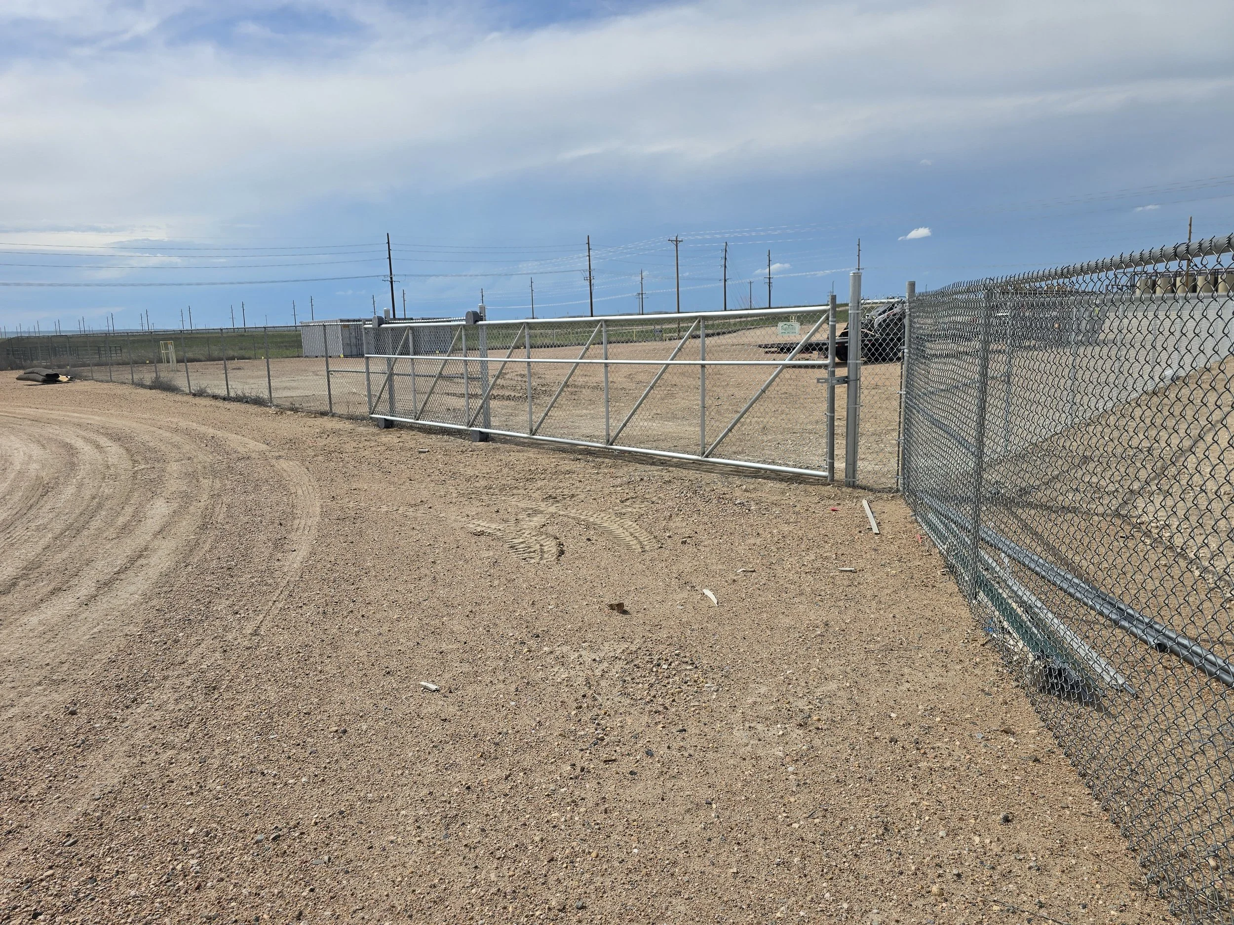 A chain-link fence with a sliding gate runs along a dirt road in a rural area, with electric poles and a car in the background under a partly cloudy sky.