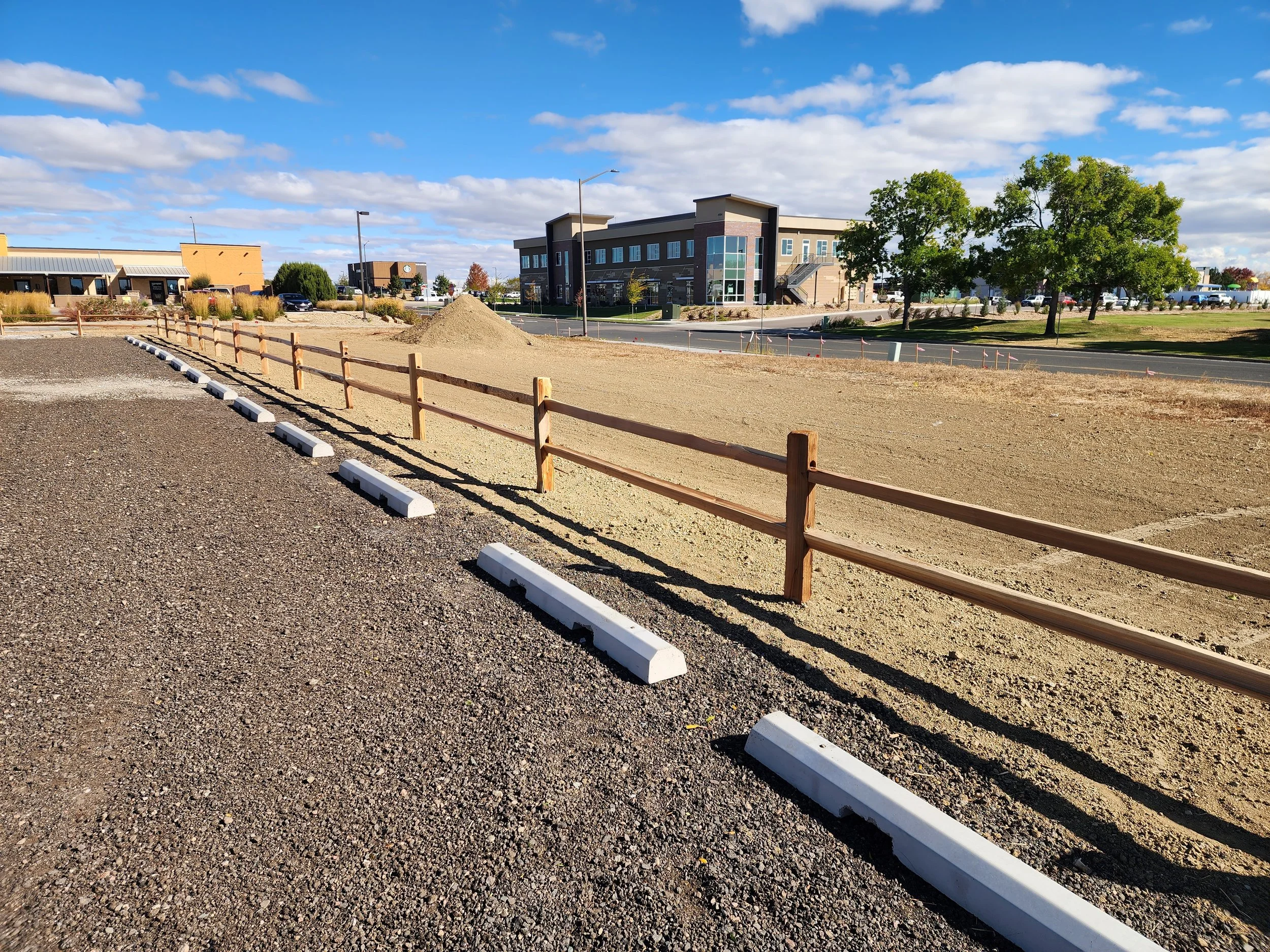 A construction area with a gravel parking lot, a wooden fence, and a pile of dirt, with a large modern building, trees, and a blue sky in the background.