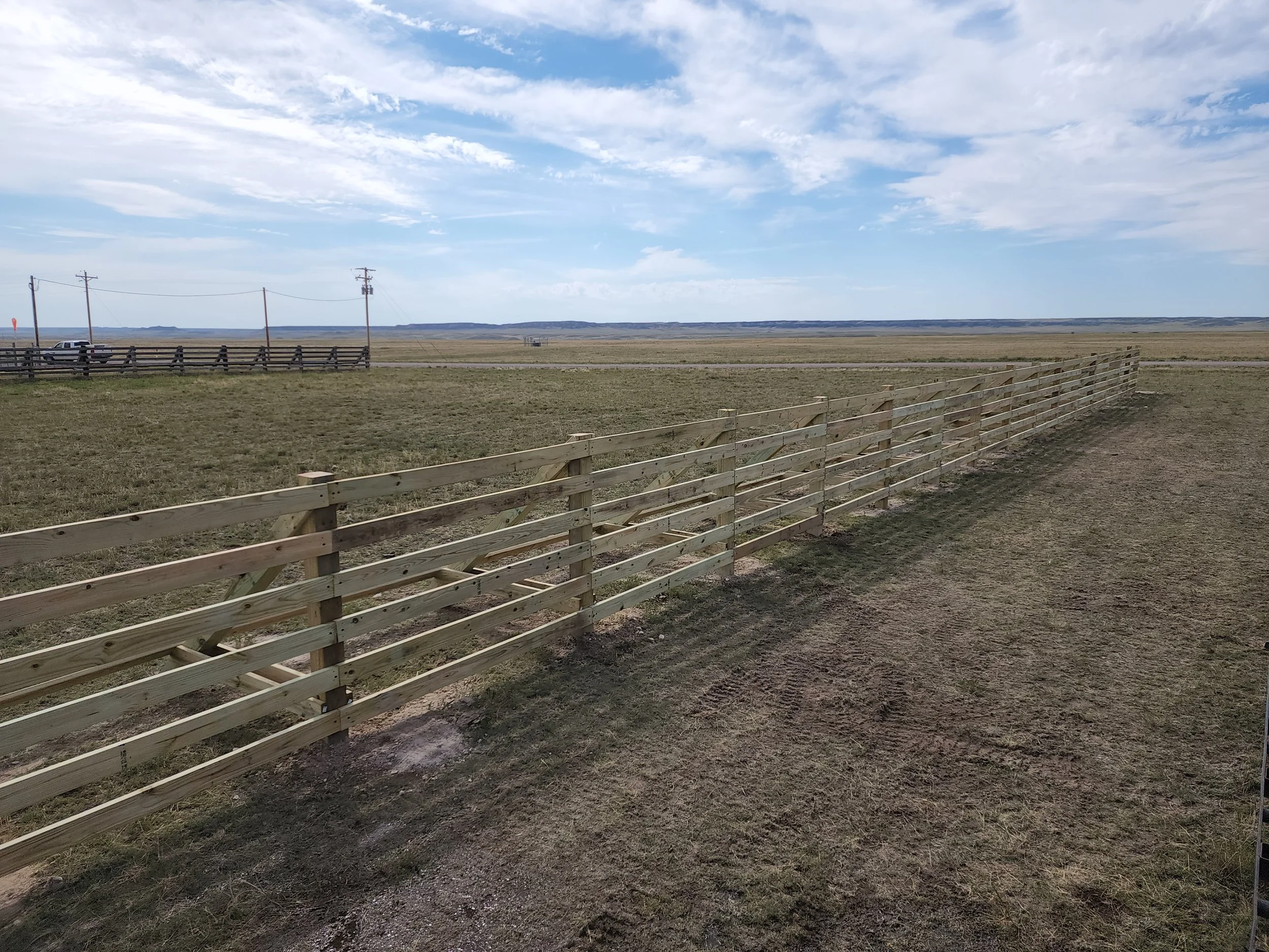 A landscape scene with a long wooden fence stretching across a grassy field, utility poles and power lines in the distance under a partly cloudy sky.