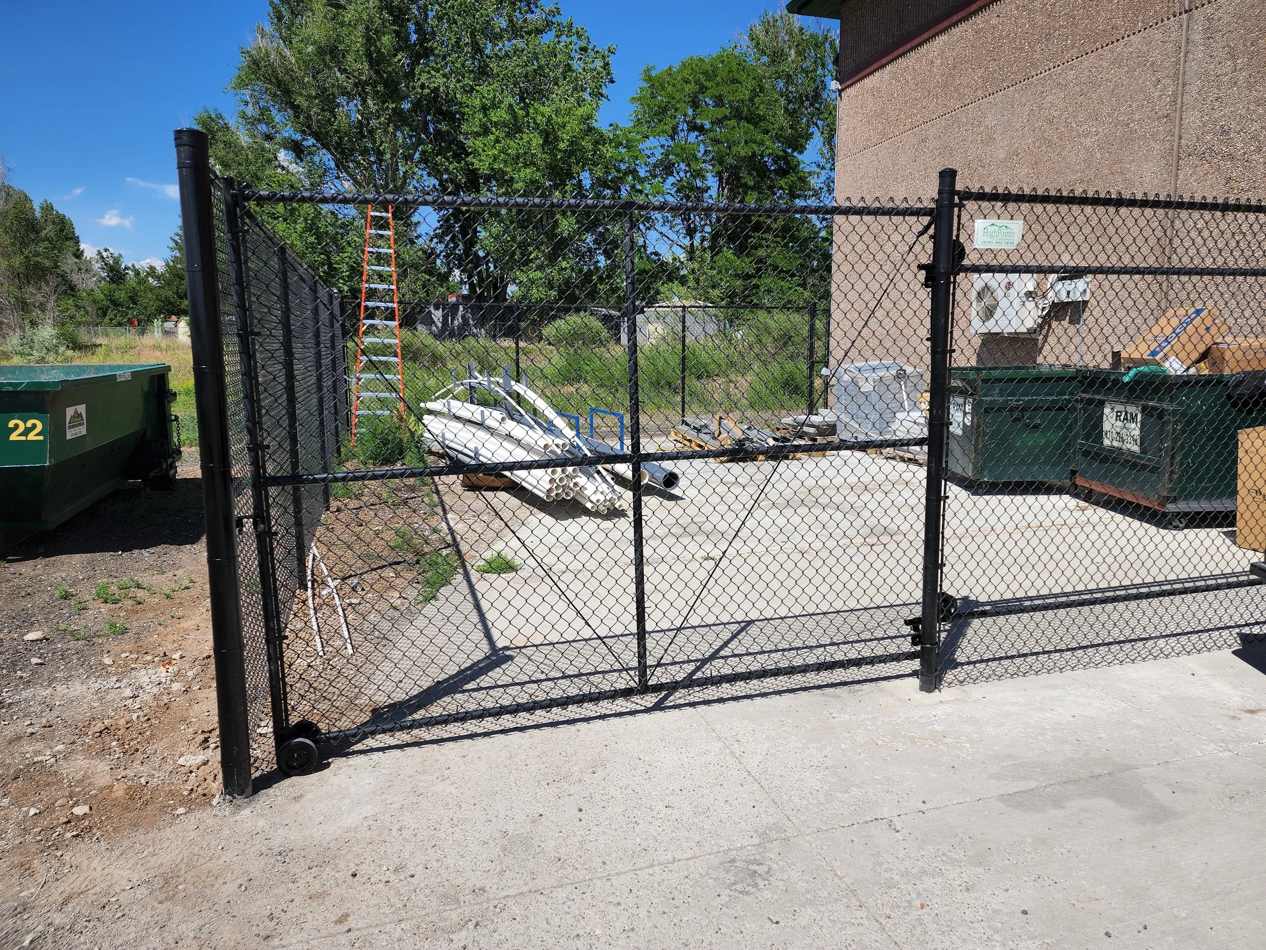 A black chain-link fence gate with a solid latch, enclosing a small outdoor area with various items including a kayak, ladder, trash cans, and construction supplies, next to a brick building and a concrete sidewalk.