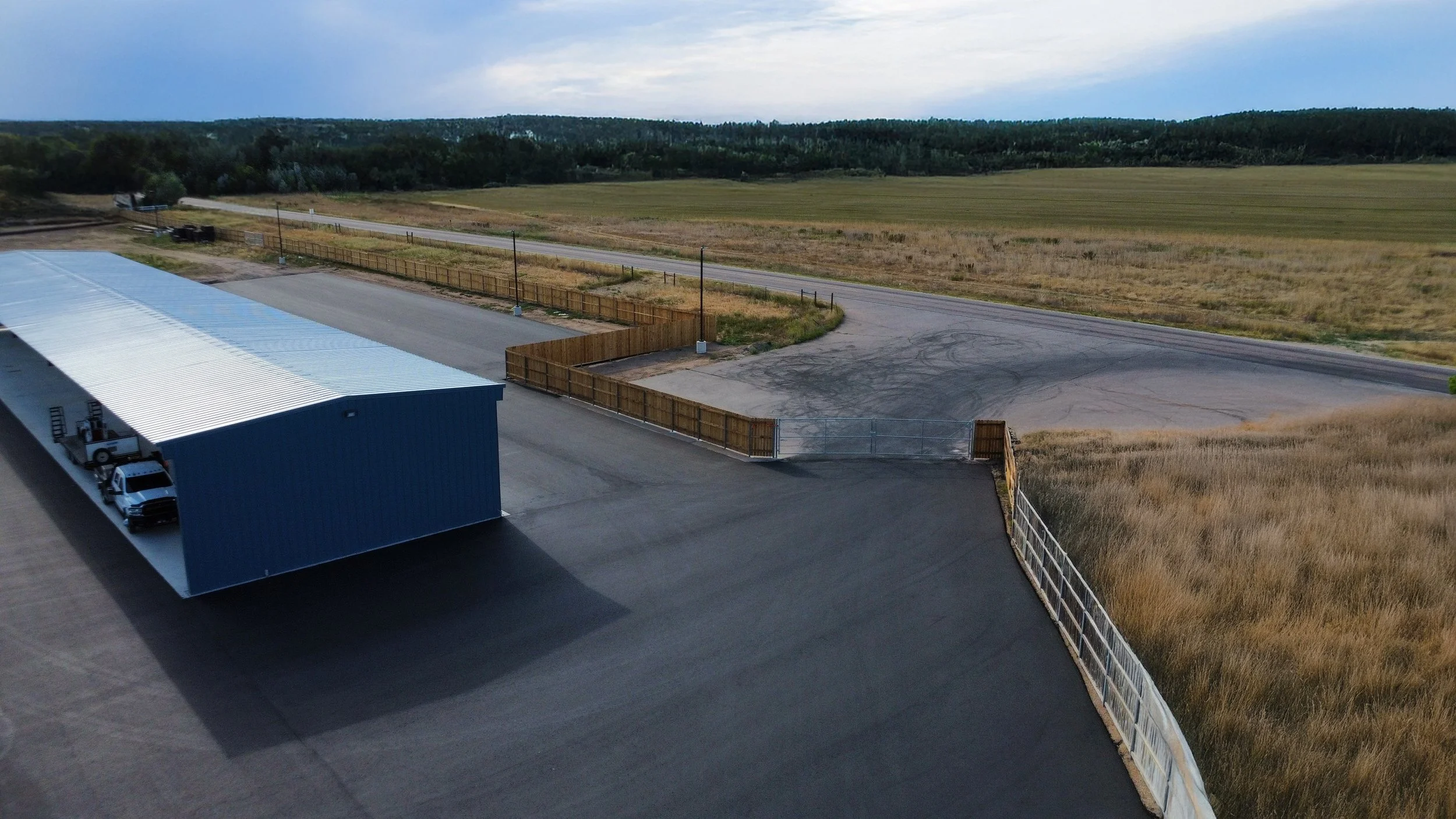 An outdoor storage area with a large blue metal building, parking space, and a fenced parking lot. The background features open fields, a rural road, and a tree-lined horizon under a partly cloudy sky.