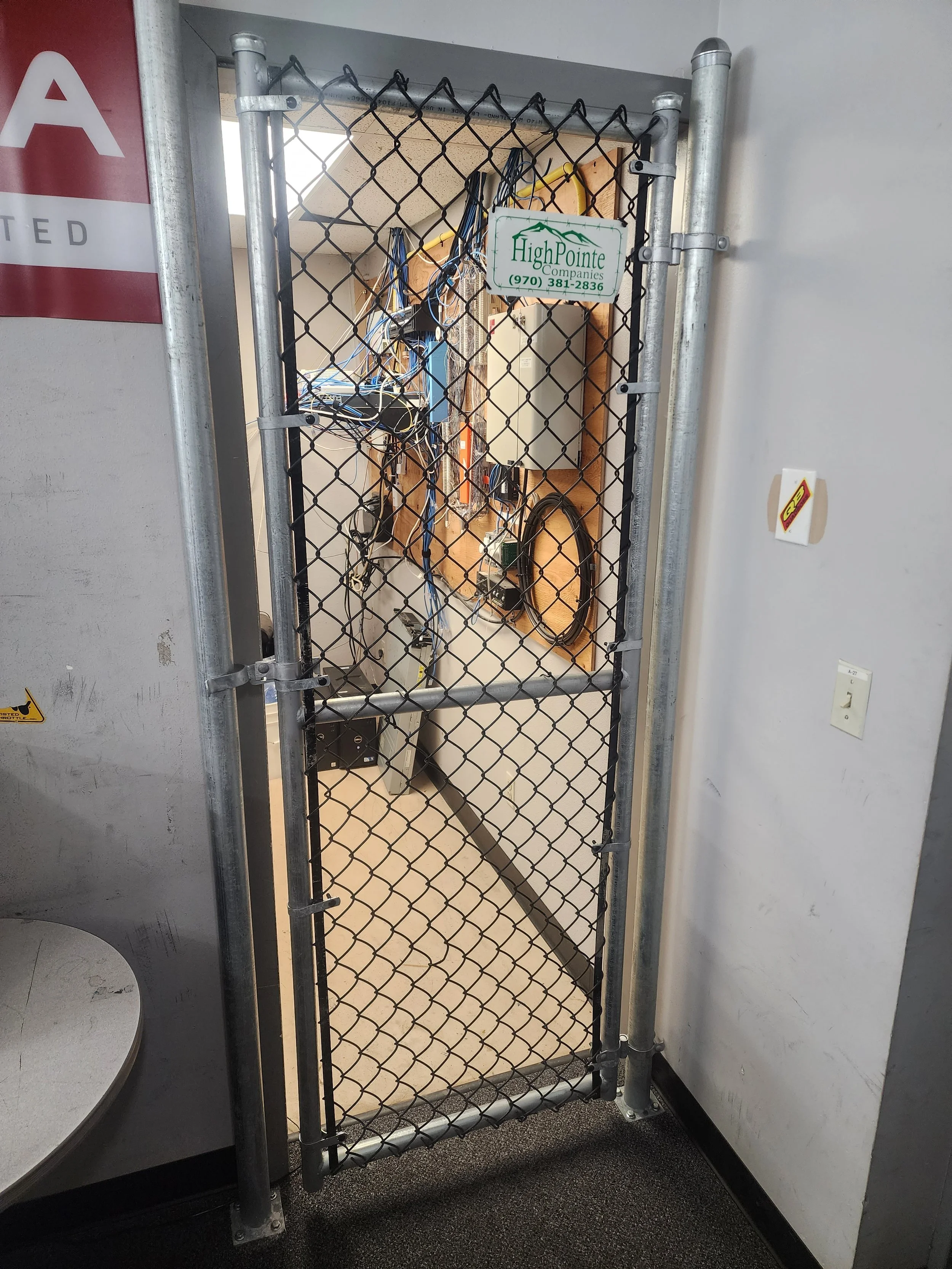A metal security gate with a lock, blocking access to a tech workspace with routers, cables, and electronic devices on a wooden wall panel.