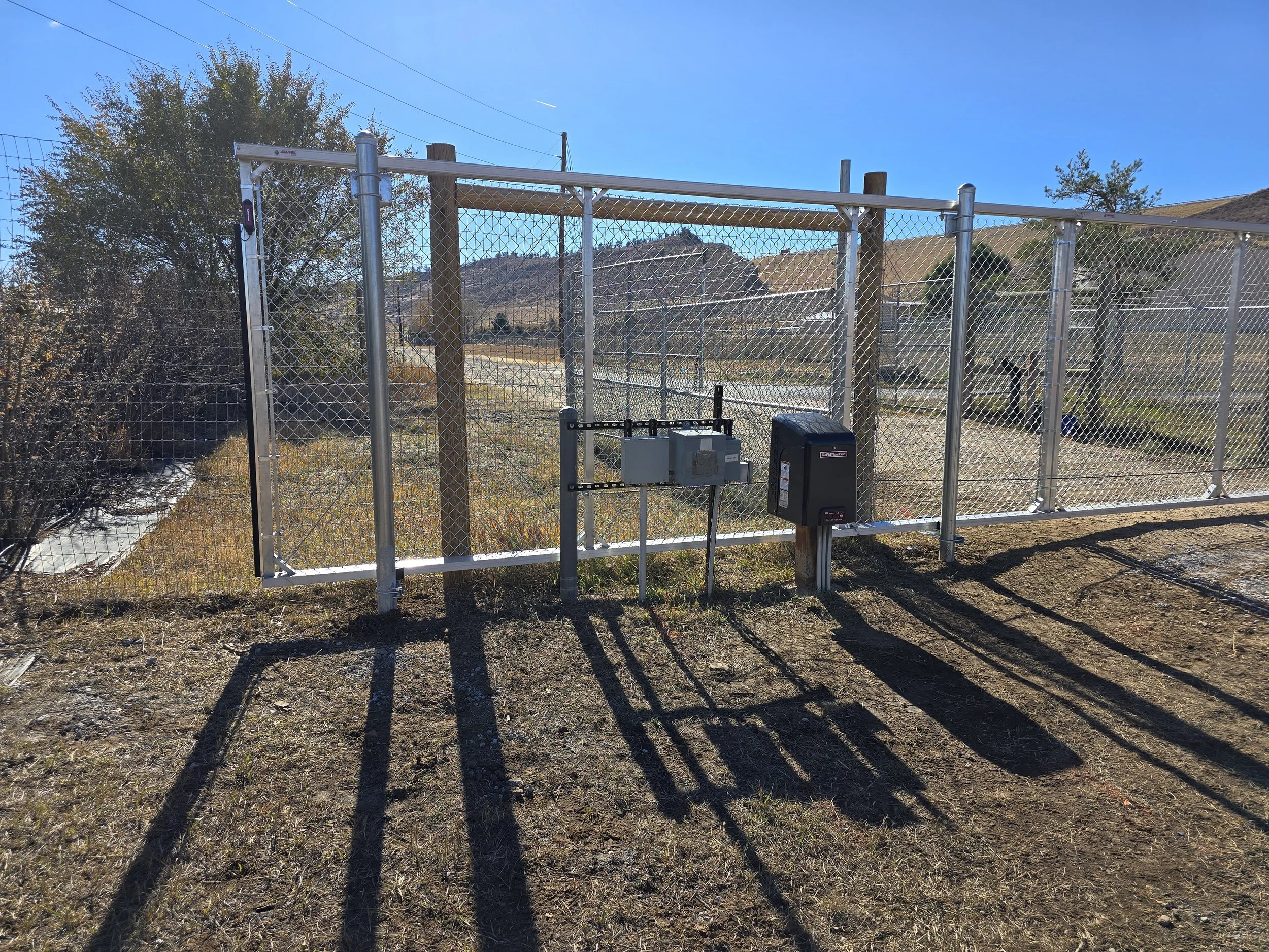 View of a fenced area with metal and chain-link gate, electrical boxes, and a box mounted on a pole, with a dirt ground and hills in the background on a sunny day.