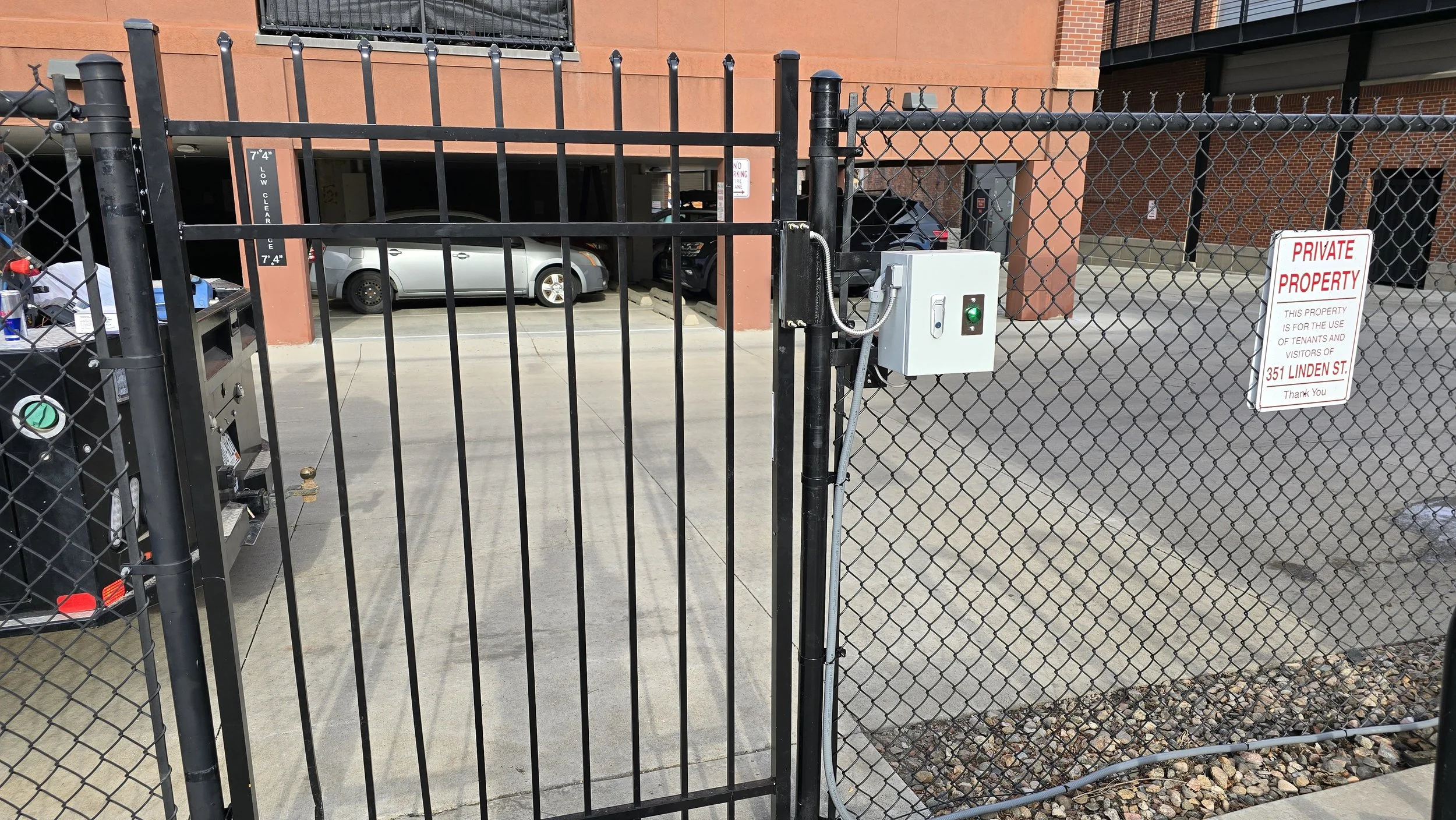 View of a parking lot gated entrance with a black metal gate and a gray chain-link fence. A sign indicates private property at 351 Linden Street. Parking spaces with cars are visible behind the gate.