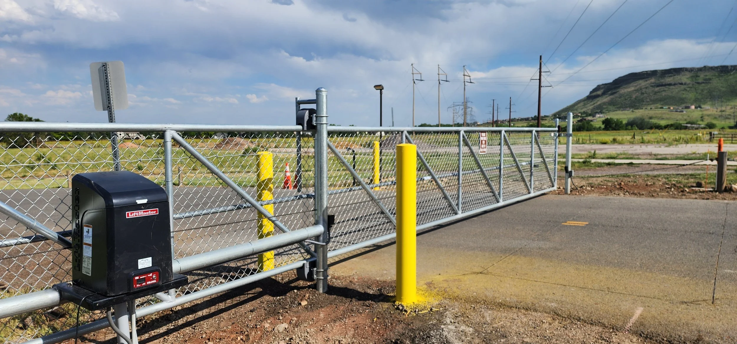 A secure metal gate with a LiftMaster automation system, yellow safety posts, chain link fencing, and a landscape with a hill and electrical poles in the background.