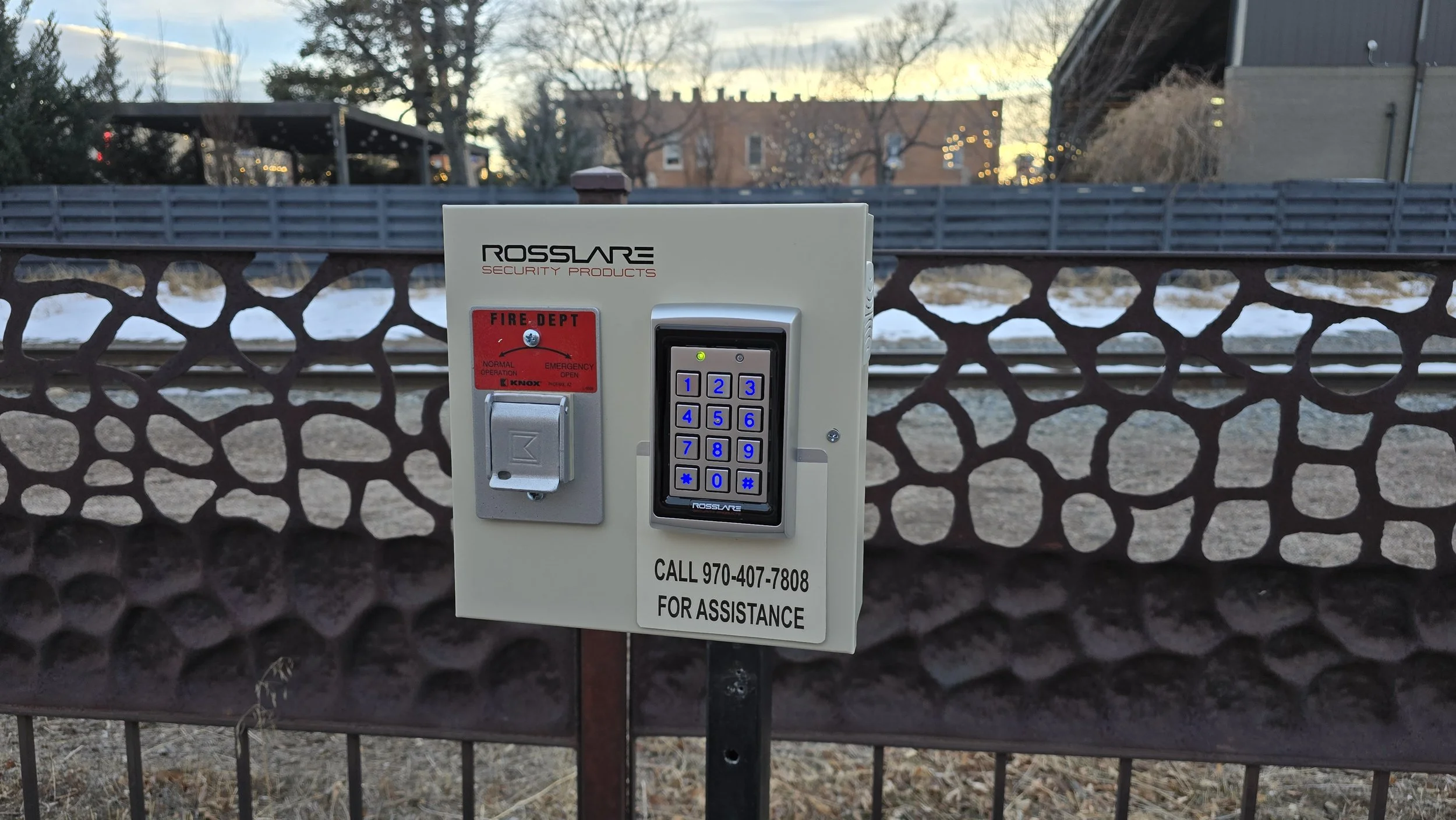 A security keypad and fire alarm box mounted on a metal post in front of a decorative metal fence with trees and buildings in the background during dusk.