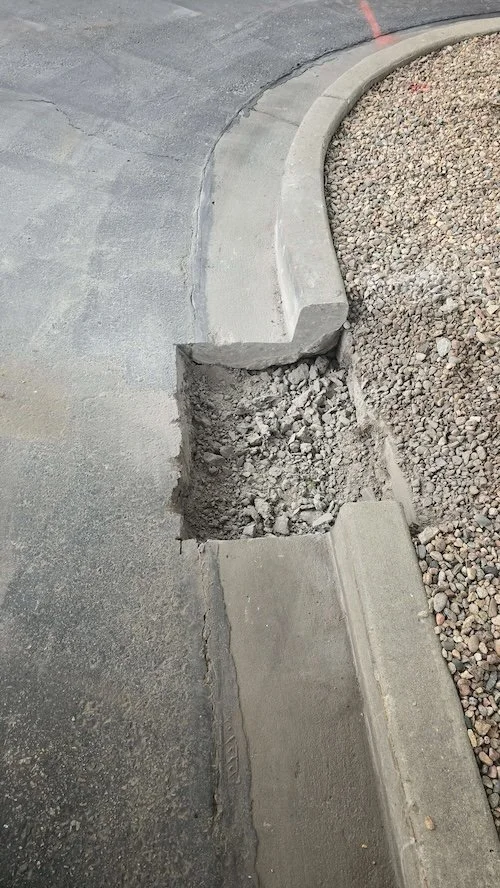 A curb with a section of concrete broken away, revealing gravel underneath, next to an asphalt road and a gravel area.
