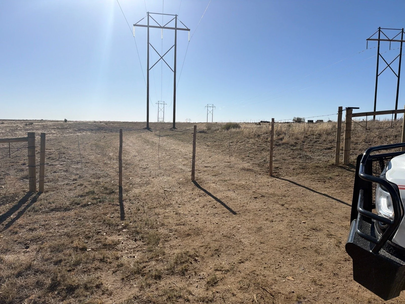 A dirt pathway leading to power transmission lines in a dry, open field under a clear blue sky, with part of a vehicle visible in the lower right corner.