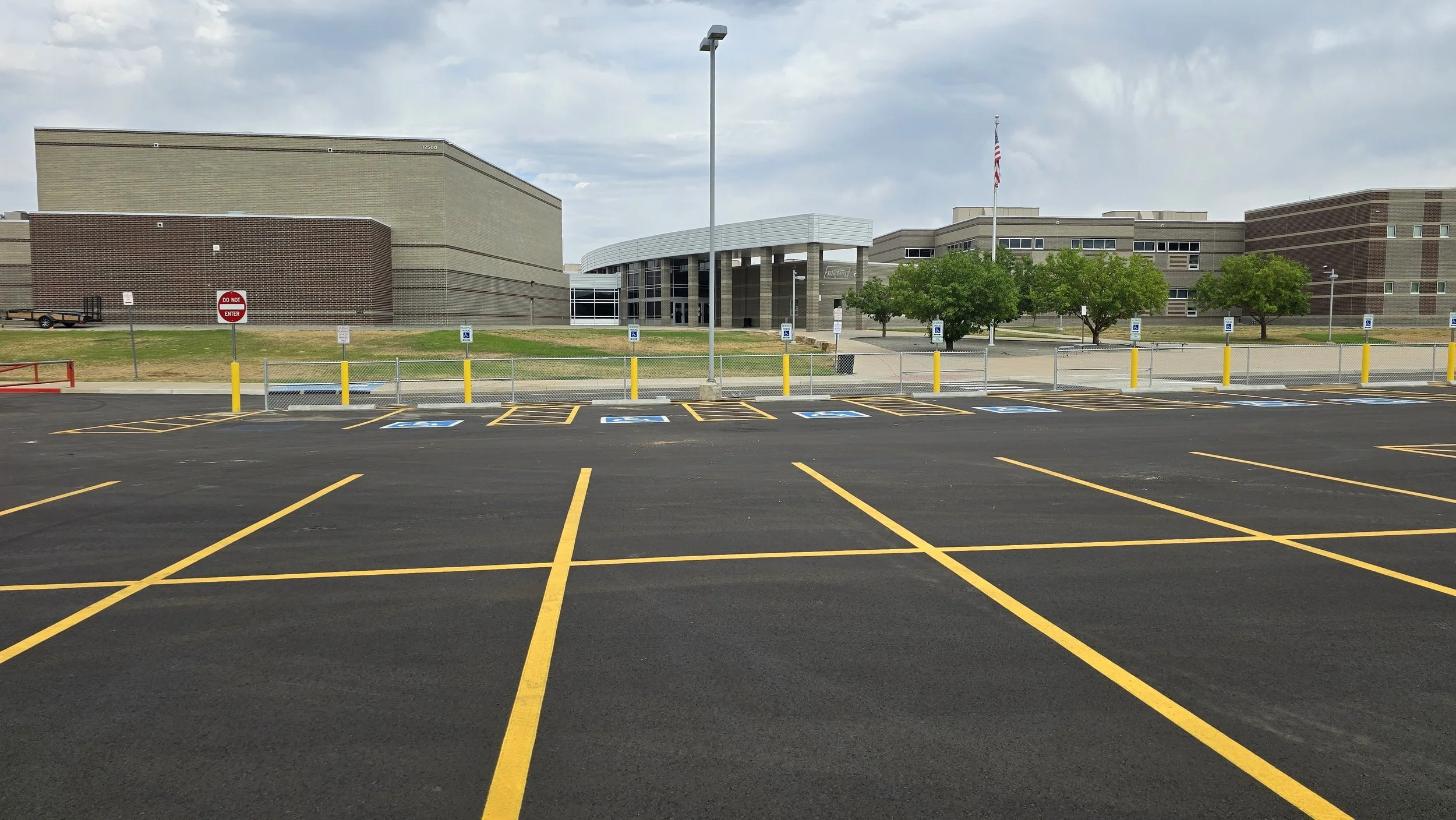 Empty parking lot with designated handicapped parking spaces in front of a school building with trees, a flag, and a cloudy sky.