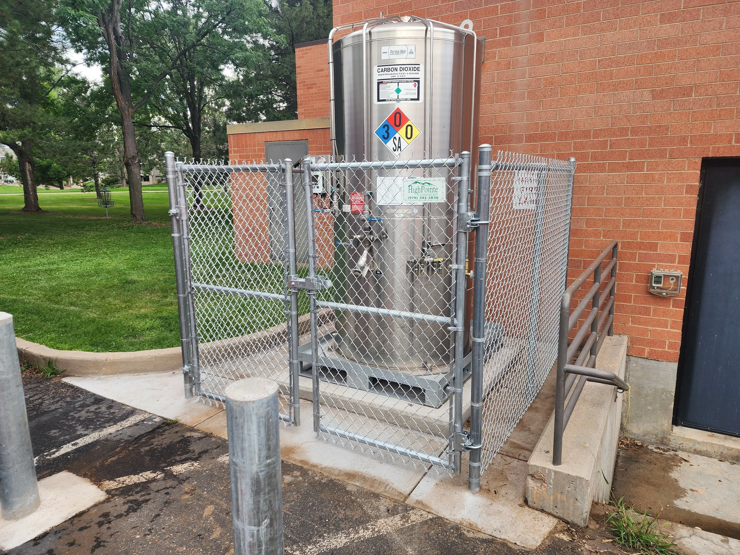Industrial tank labeled 'Carbon Dioxide' enclosed in a metal fence outside a brick building with green grass and trees in the background.