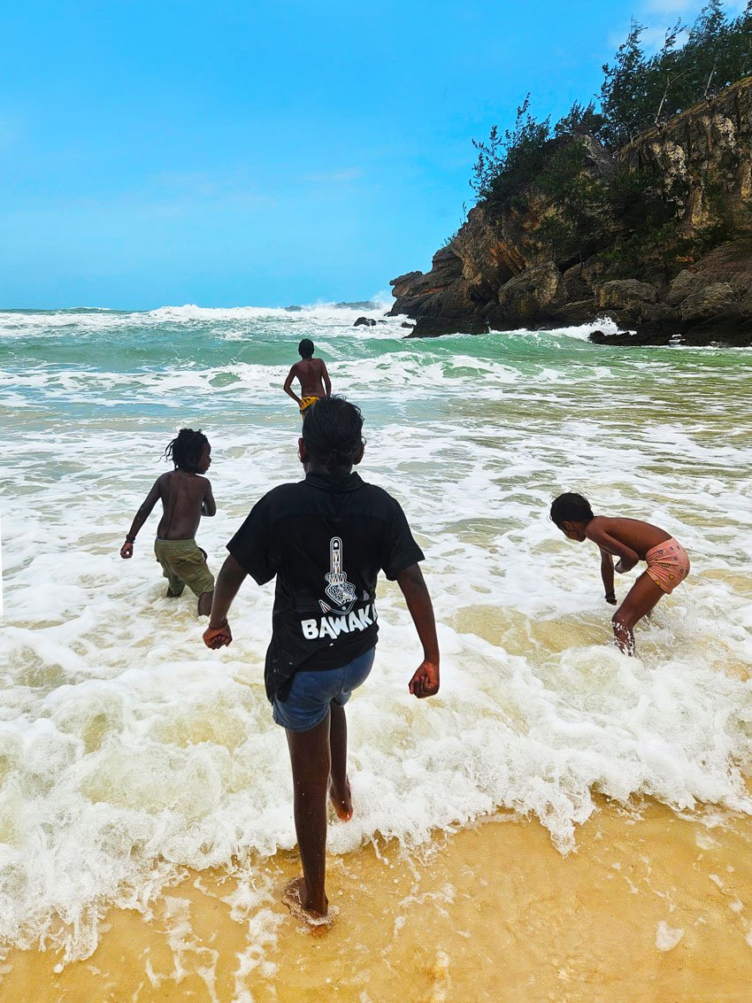 Four children playing at the beach near a rocky shoreline under a blue sky.
