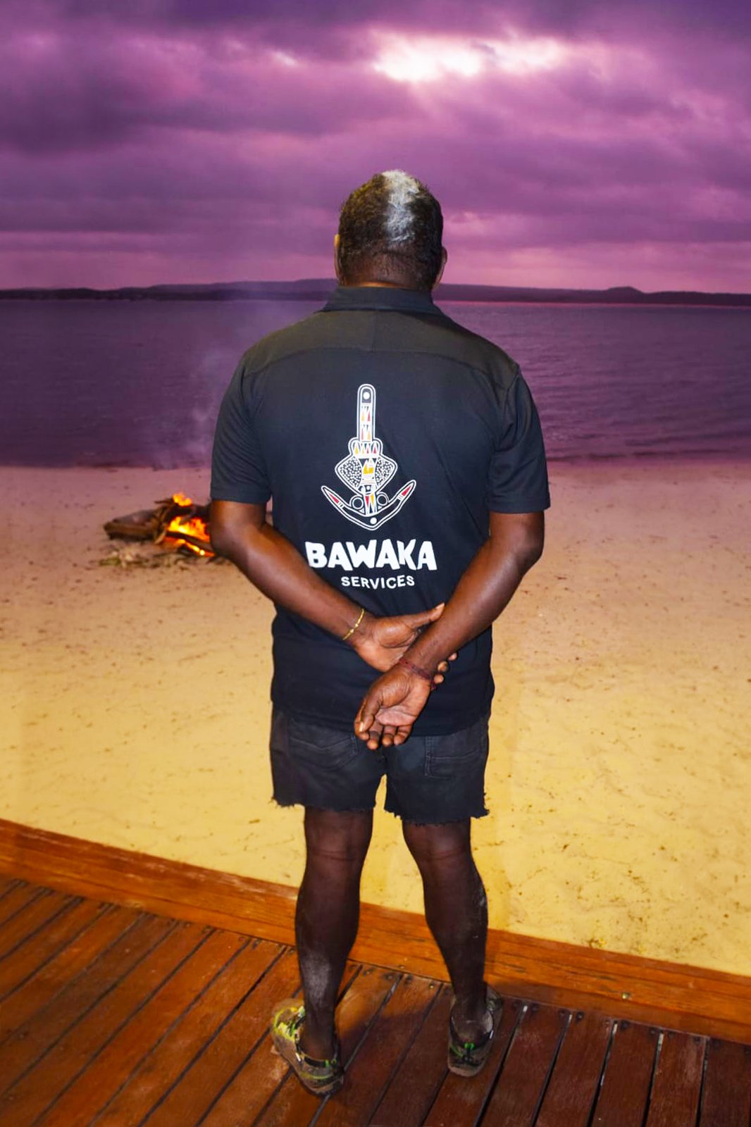 Man standing on wooden deck facing a beach and lake at sunset or dusk, wearing a black T-shirt with 'BAWAKA SERVICES' and a logo on the back, with purple clouds in the sky and a small campfire on the sand to the left.