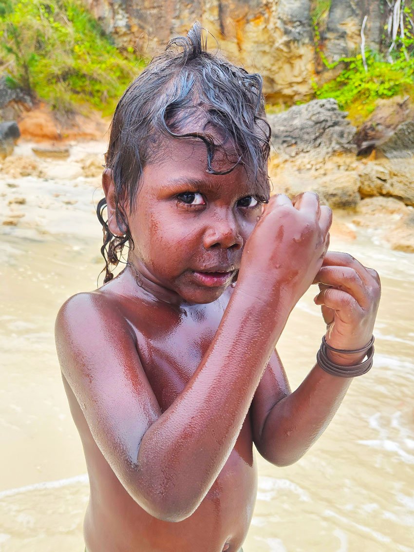 Young boy with wet hair and dark skin standing in a sandy beach with rocky cliffs in the background, holding his hand close to his face while looking at the camera.