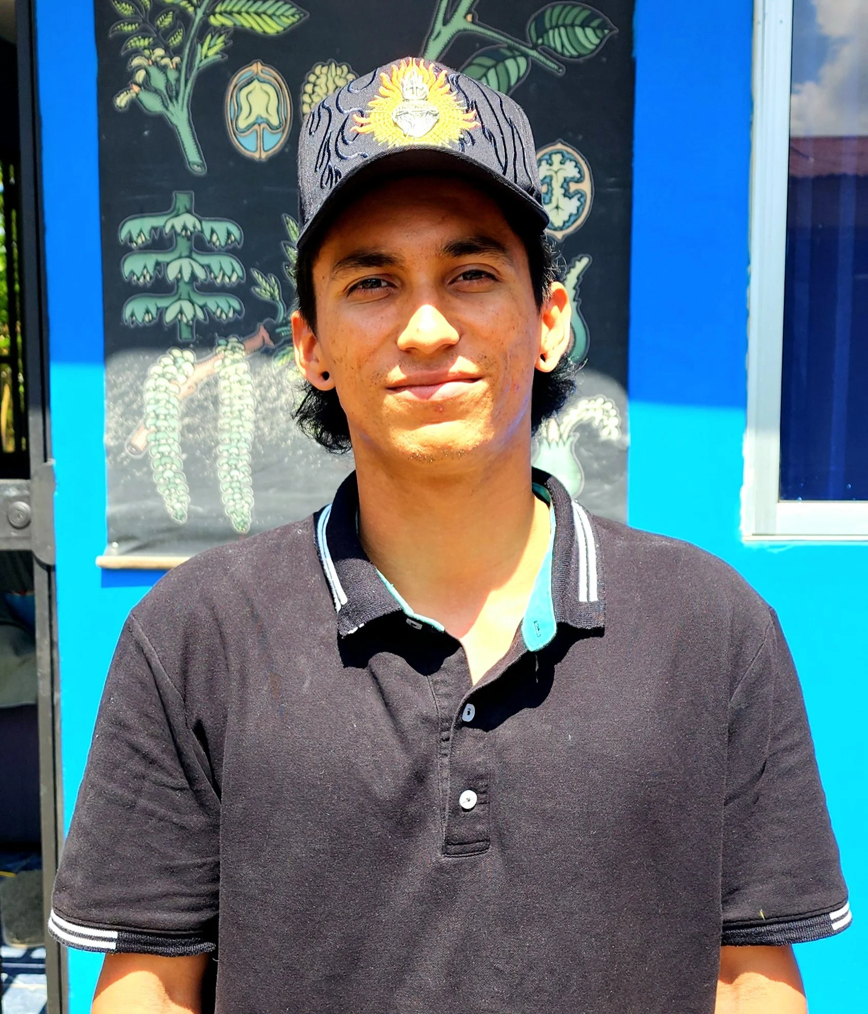 A young man with dark hair, wearing a black polo shirt and a black cap with yellow and purple embroidery, stands in front of a colorful botanical poster on a blue wall.