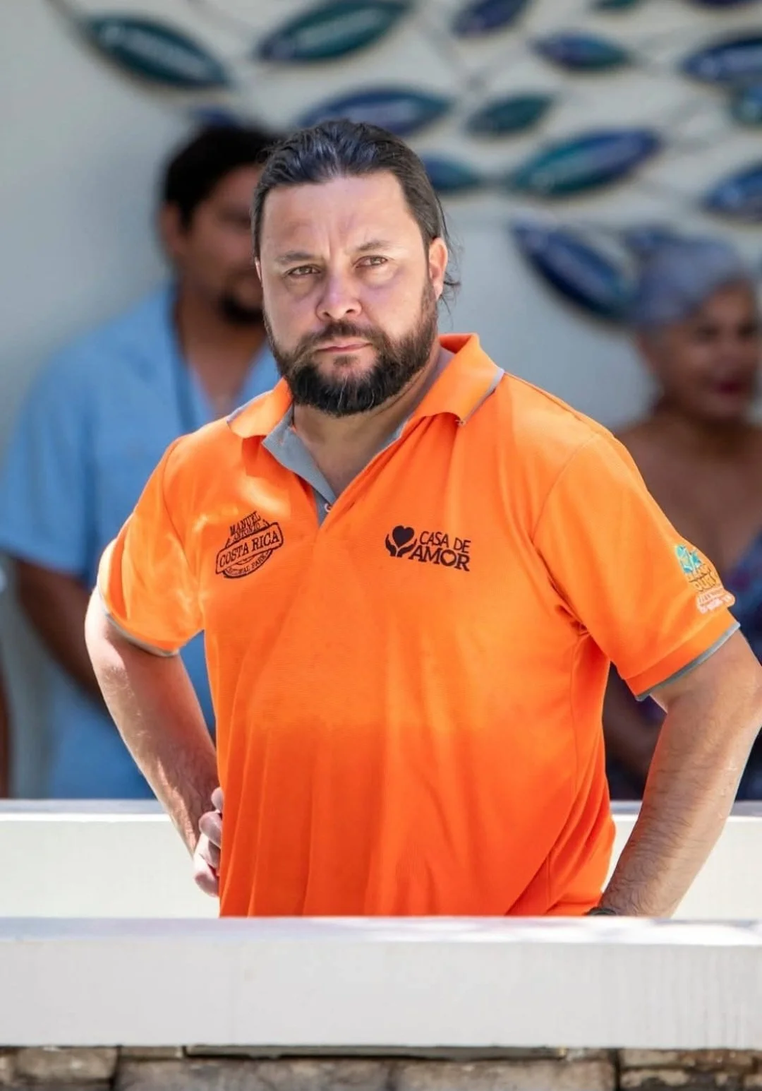 A man with a beard and long dark hair, wearing an orange polo shirt, standing outdoors with his hands on his hips, with other people in the background.