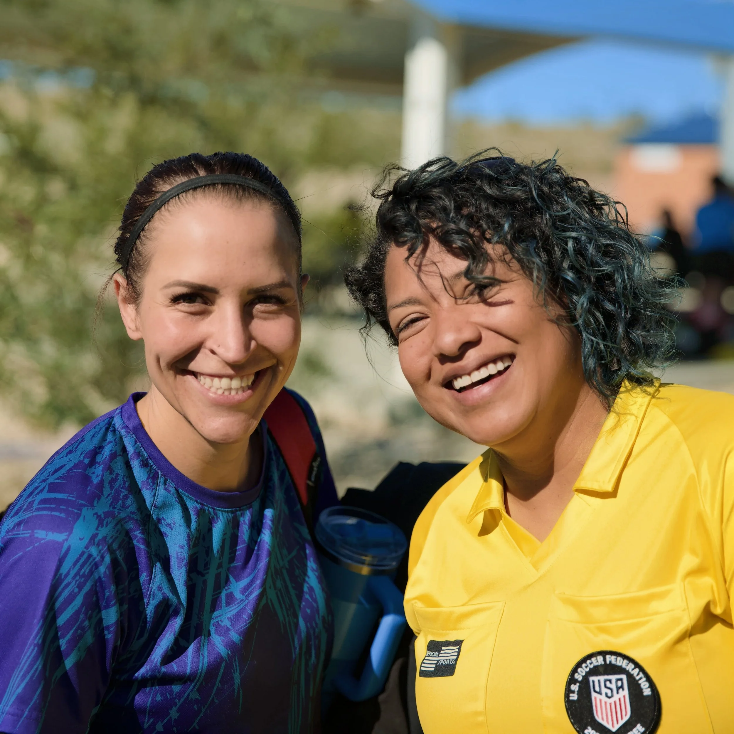 Two women smiling outdoors, one wearing a purple sports shirt and the other in a yellow soccer jersey with USA Soccer Federation patch.