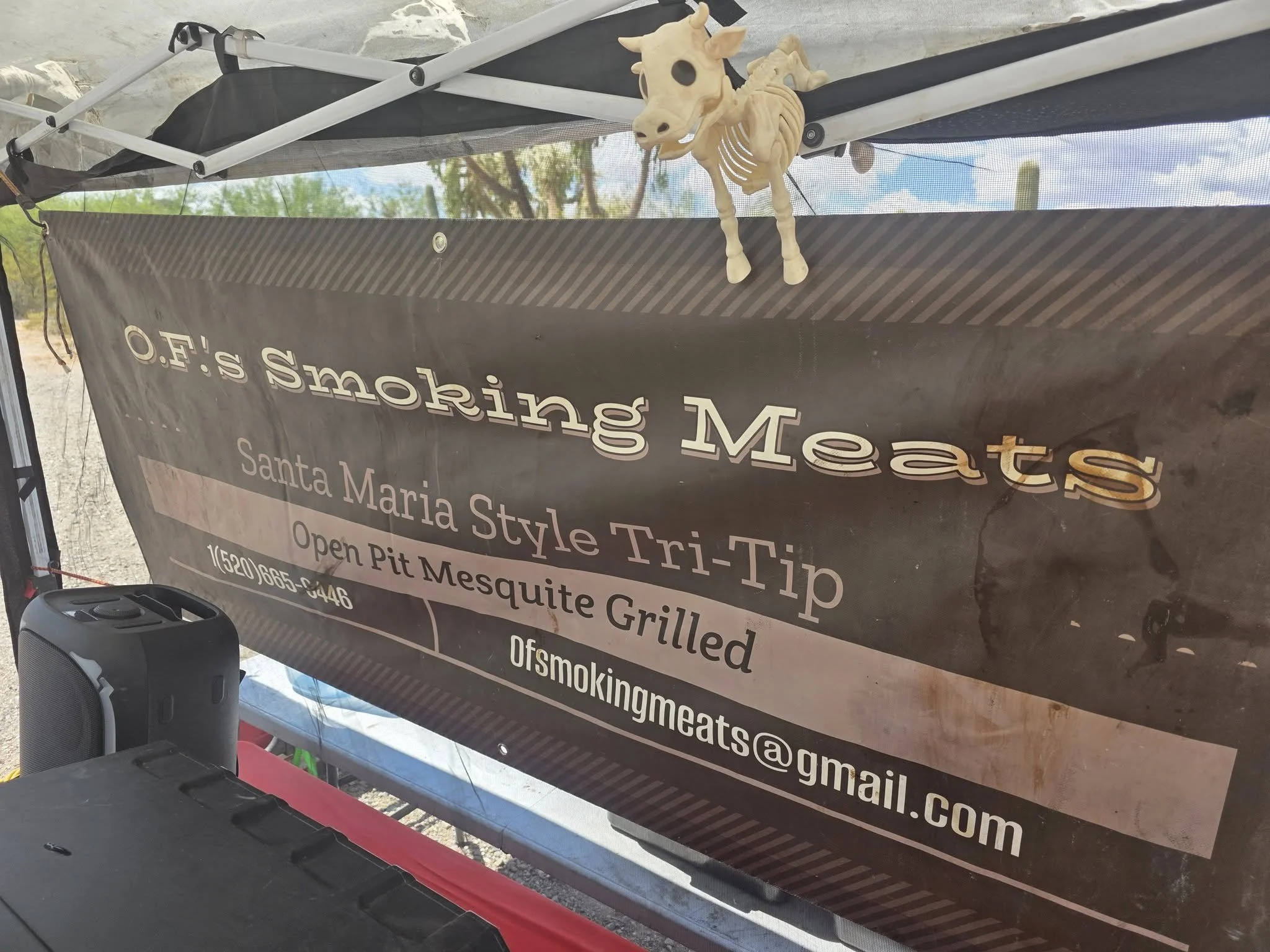 A black banner hanging under a tent advertising Smoking Meats, Santa Maria Style Tri-Tip, open pit mesquite grilled, with a skeleton decoration attached to the tent's frame.
