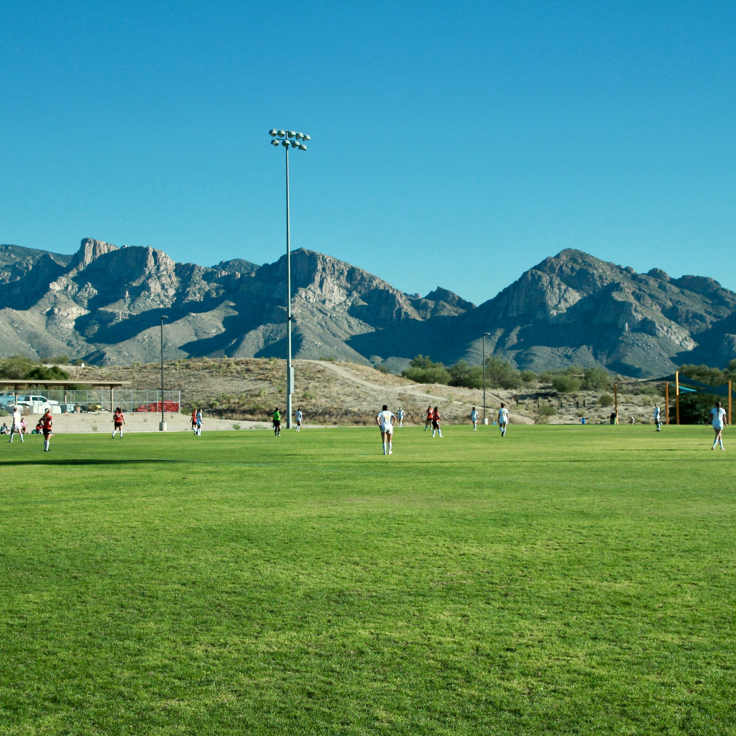 A soccer game is being played on a grassy field with mountains in the background and a clear blue sky.
