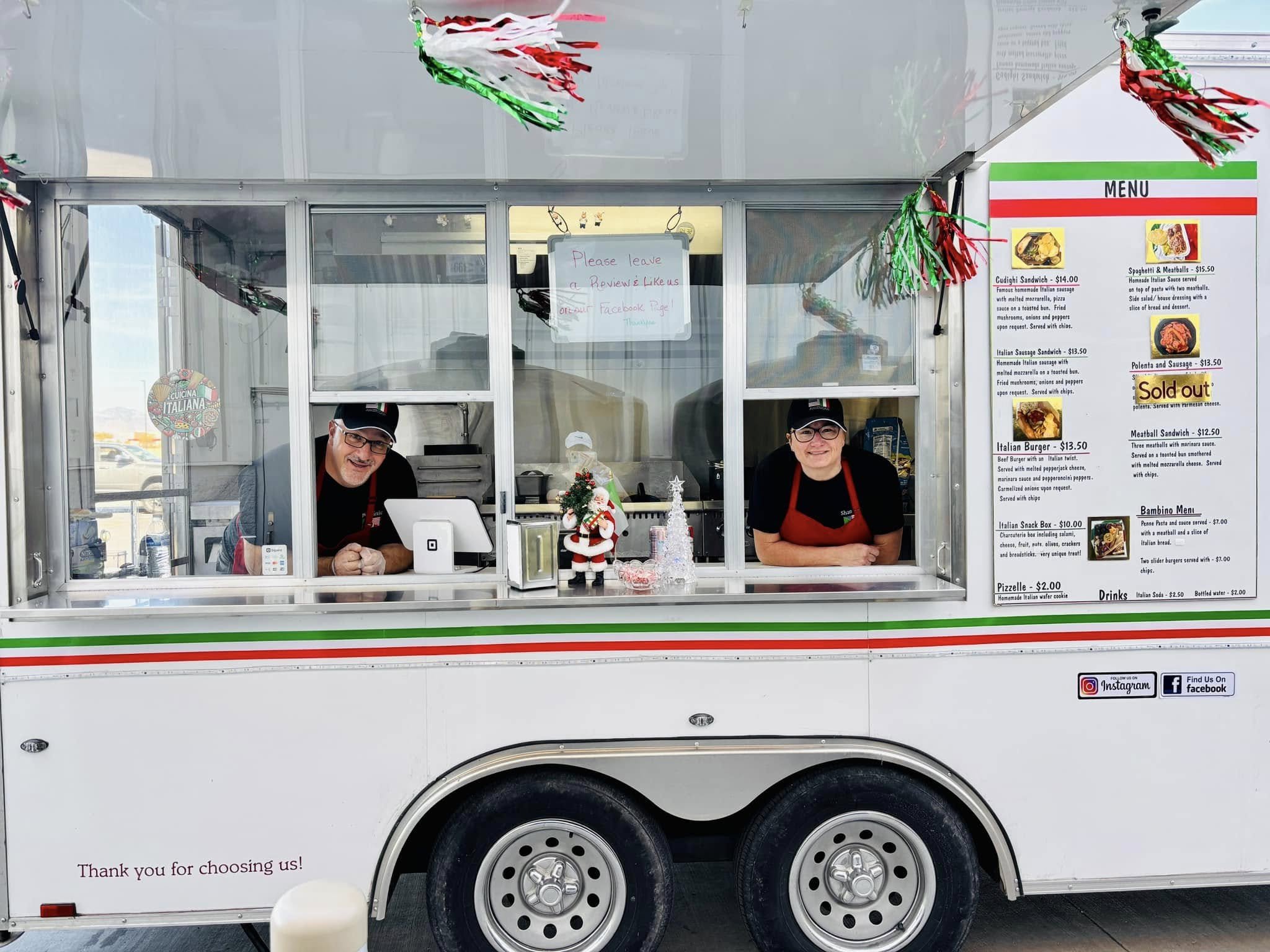 A food truck decorated for Christmas with two workers inside, a man and a woman, both wearing hats and red aprons. The truck has a Christmas ornament, a small Christmas tree, and a snowman decoration on the counter. The menu board lists various Itali