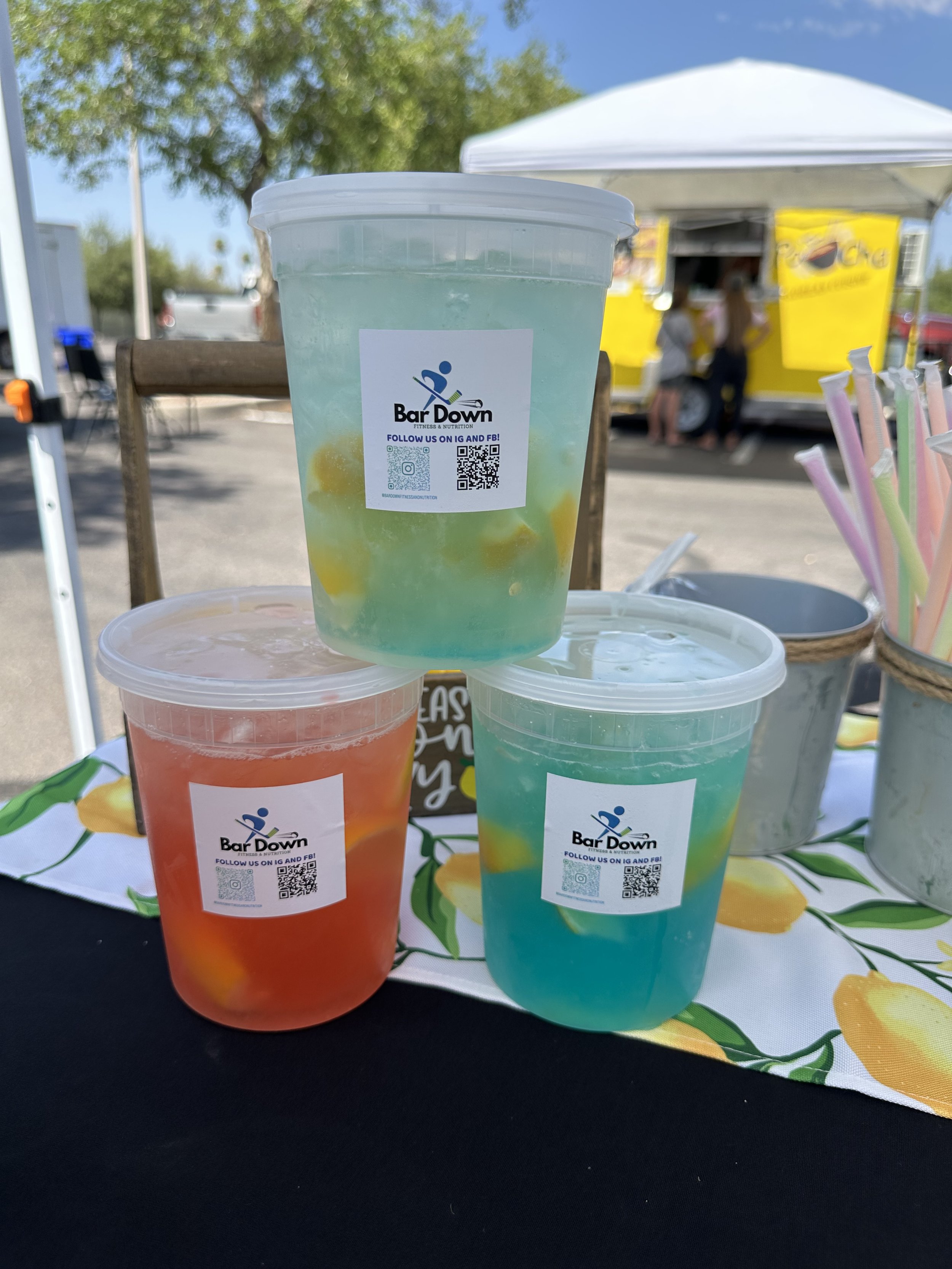 Three colorful drinks in clear cups with a sticker that says 'Bar Down' and QR codes, set on a table with a lemon-patterned cloth at an outdoor event with a food truck and tents in the background.