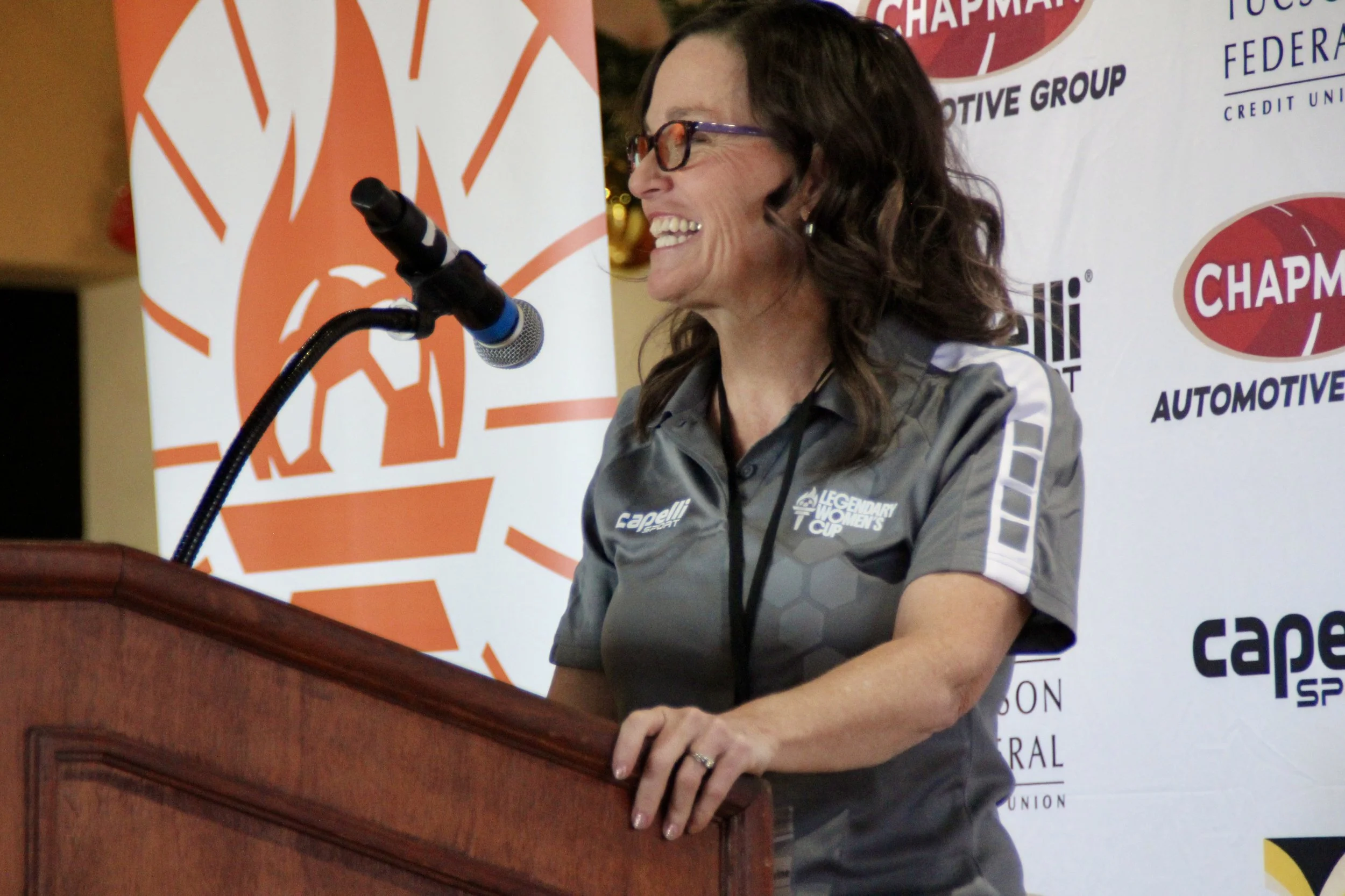 A woman with brown hair and glasses smiling while speaking at a podium, wearing a gray sports shirt with logos. Behind her is a backdrop with various sponsor logos, including 'Legendary Women's Cup' and 'Capelli Sports.'