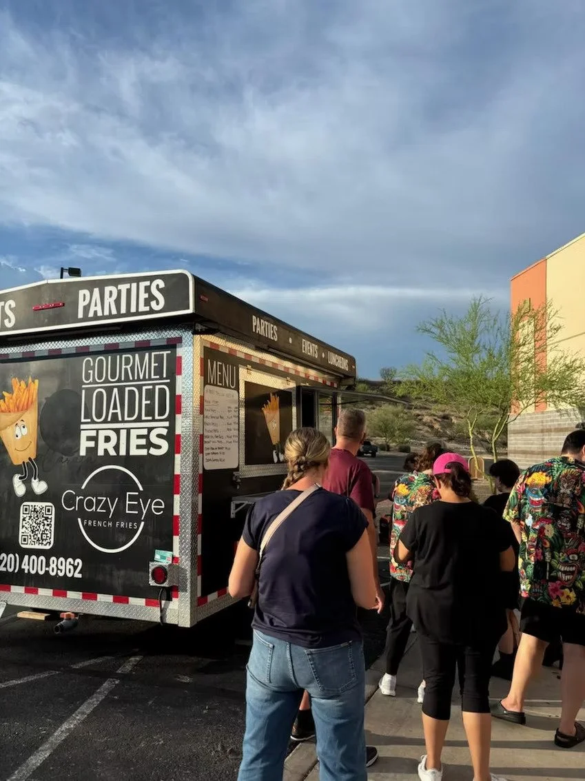 People standing in line at a food truck called Crazy Eye that offers loaded fries and other menu items in an outdoor parking lot, with a partly cloudy sky and some trees in the background.