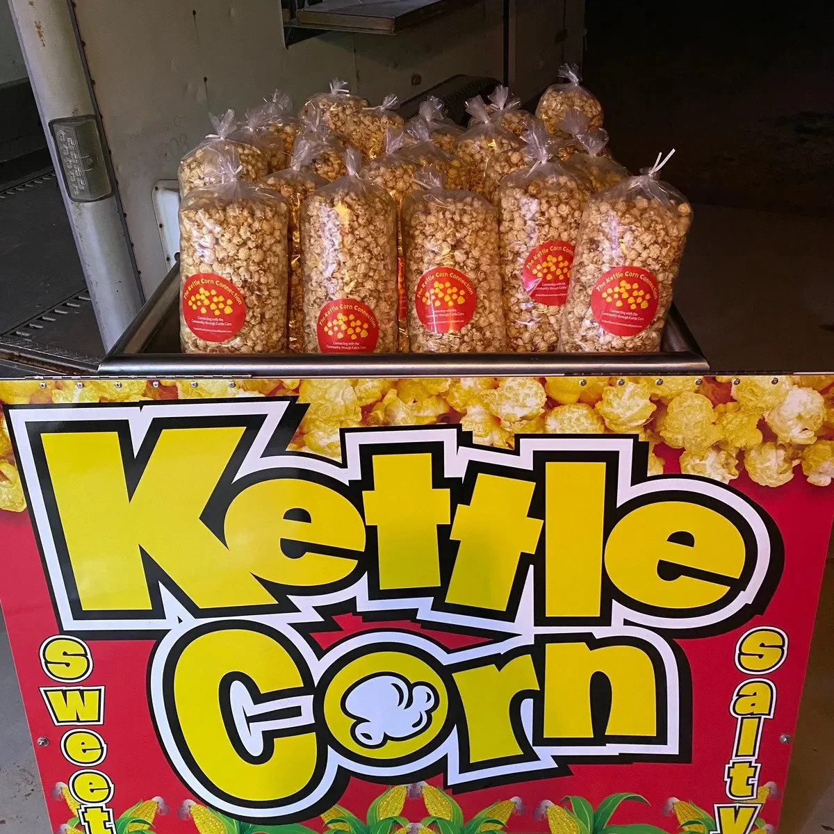 A popcorn stand decorated with a colorful sign that says "Kettle Corn," displaying bags of caramel popcorn on top and images of popcorn and corn cobs on the front.