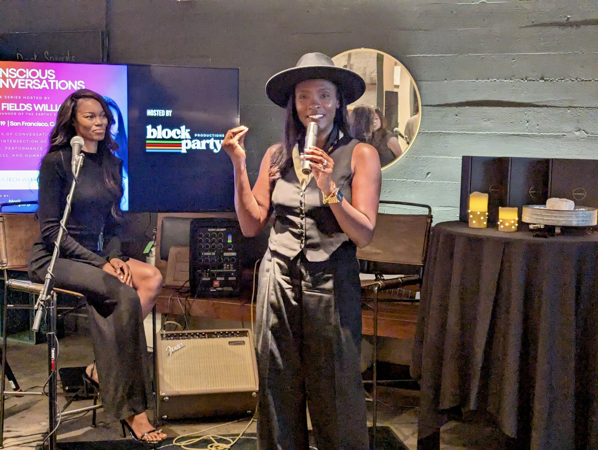 Two women speak at an event with microphones; one is standing and wearing a hat, the other is sitting. Behind them is a screen showing 'Conscious Conversations' and 'Hosted by Block Party.' The setting is indoors with professional audio equipment and candles on a table.