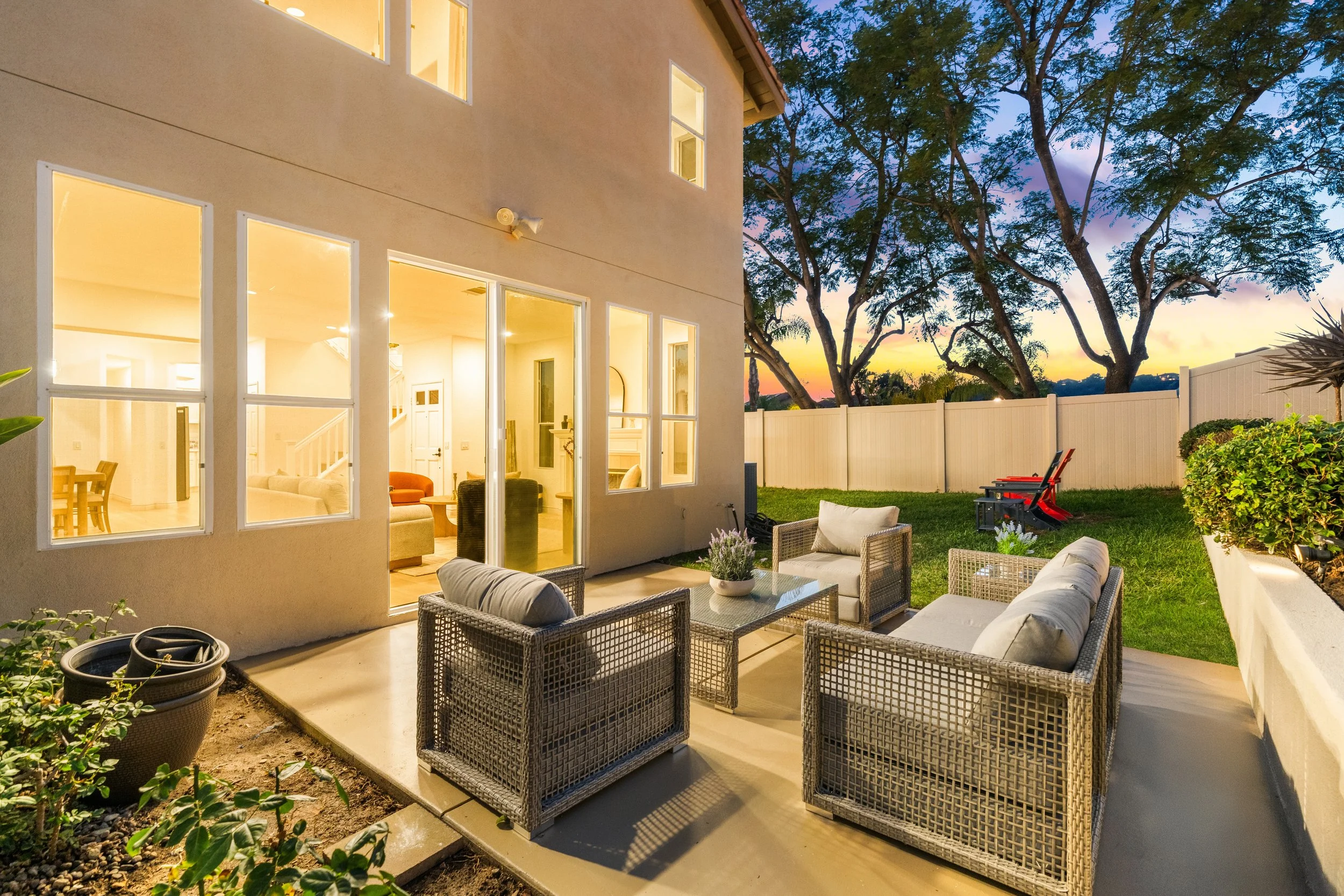 View of a house's backyard patio at sunset. The patio has wicker furniture with cushions, a glass table, potted plants, and a fenced yard with some trees and outdoor chairs.