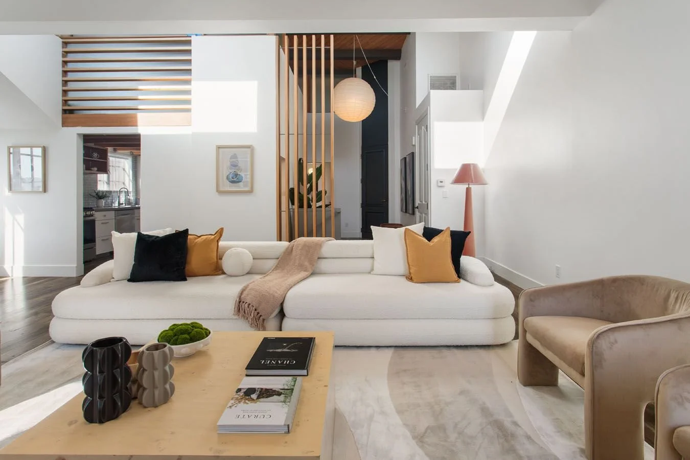 Modern living room with a white sofa decorated with black, white, and mustard pillows, and a beige throw blanket. A wooden coffee table holds black and beige vases, and magazines. A beige armchair is on the right. The room features white walls, wood accents, a paper lantern, a pink floor lamp, and artwork on the walls. The background shows a kitchen and an open entrance to another room.