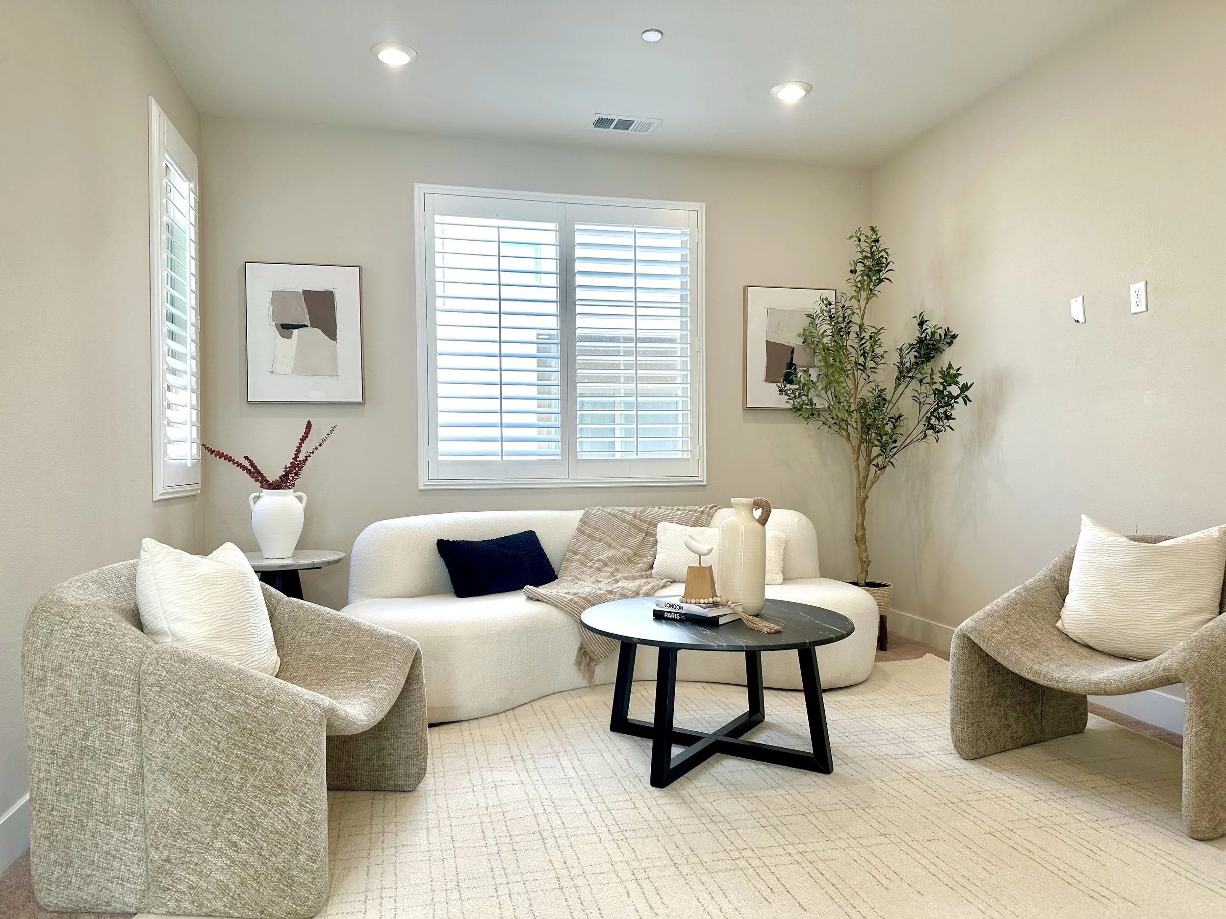 Living room with cream-colored sofa, two beige armchairs with pillows, black coffee table with decorative items, a potted tree, framed artwork on the walls, and white plantation shutters on the window.