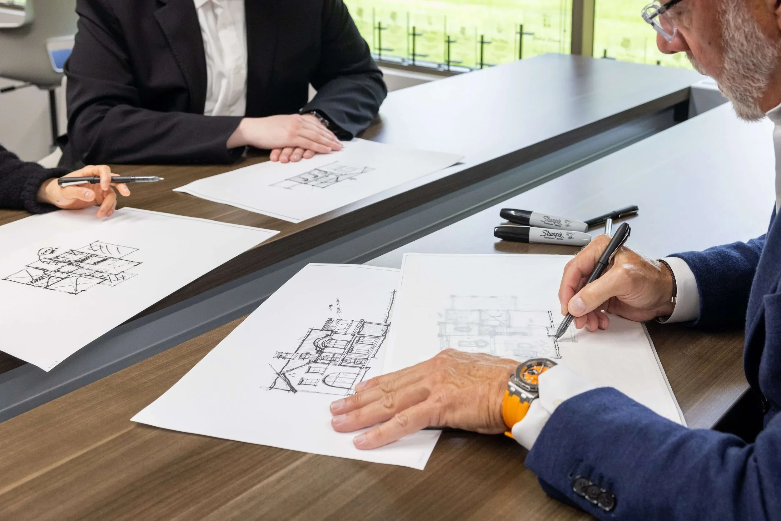 People at a meeting table reviewing architectural sketches and blueprints, with markers and pens on the table.