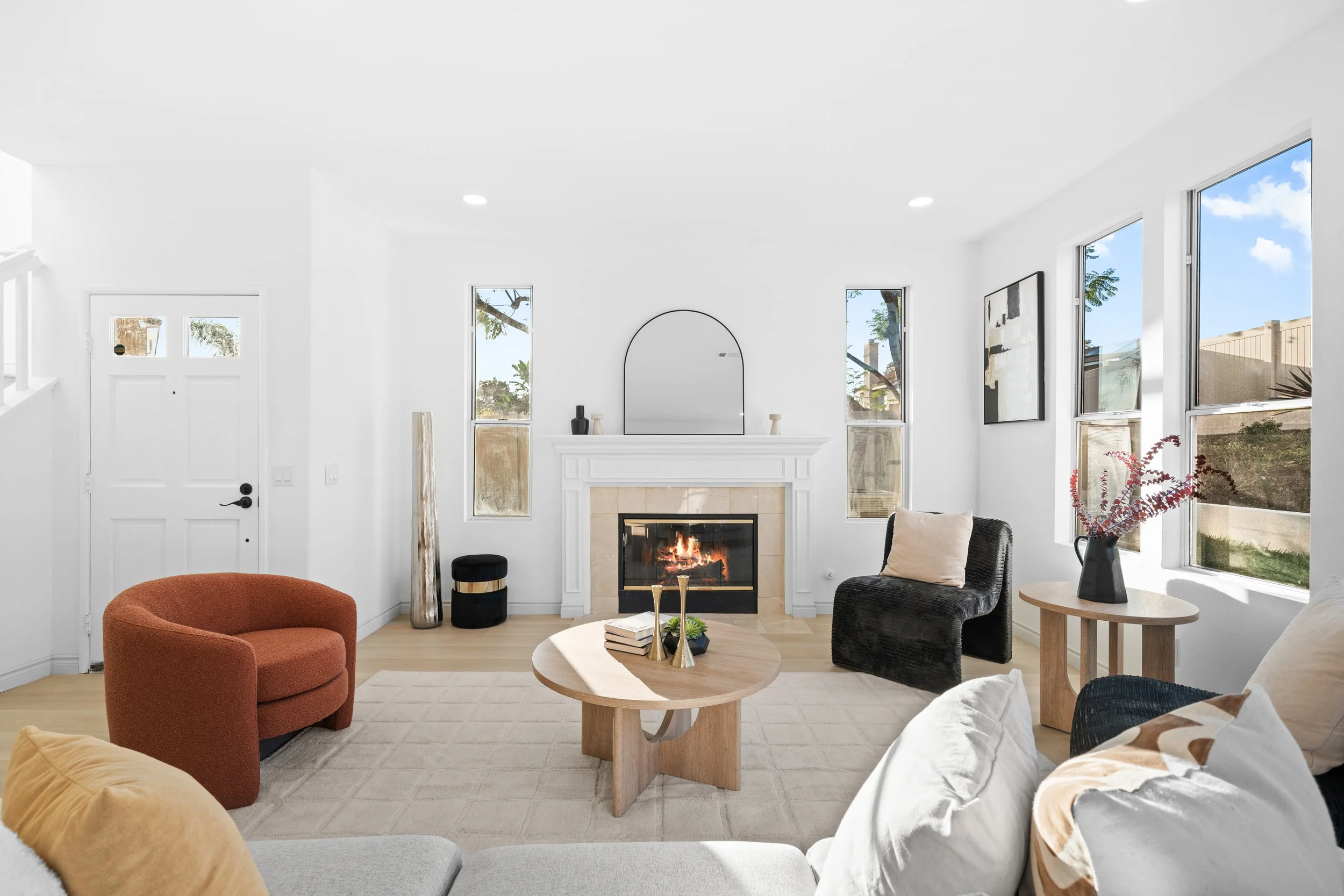 Bright living room with white walls, fireplace, and large windows showing trees and sky, decorated with modern art, furniture, and plants.