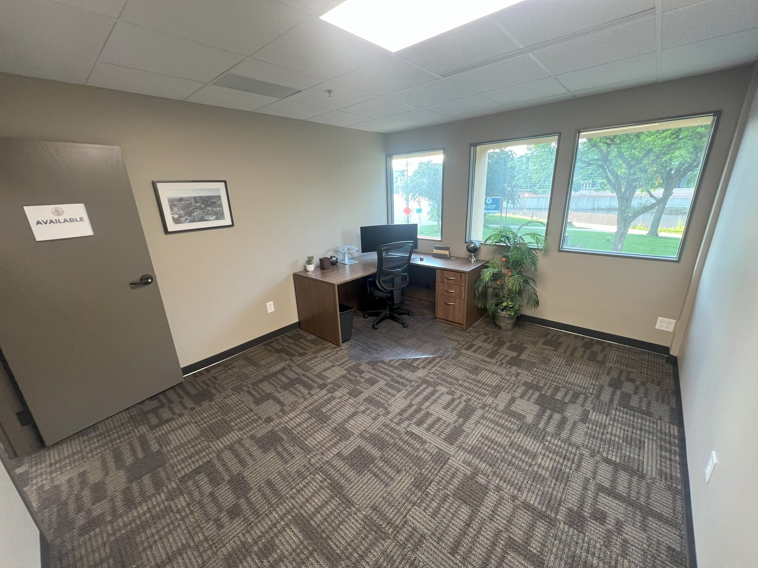 Empty office with a desk, chair, computer, and large windows showing trees outside.
