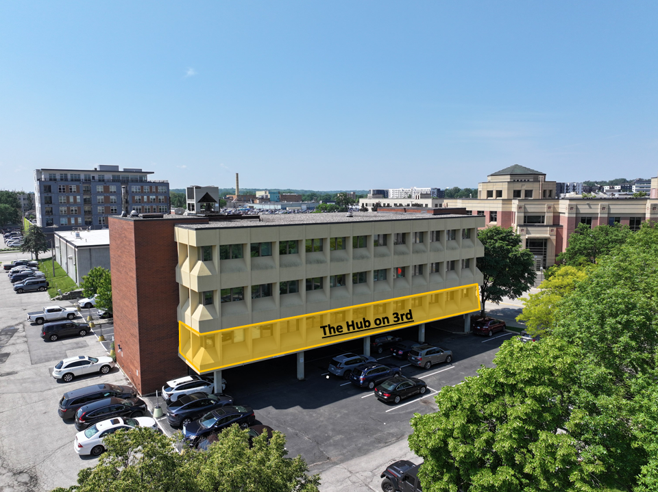 An aerial view of a multi-story building with a yellow highlighted balcony on the 3rd floor that has text reading 'The Hub on 3rd'. There are cars parked underneath and around the building, with green trees and a cityscape in the background.