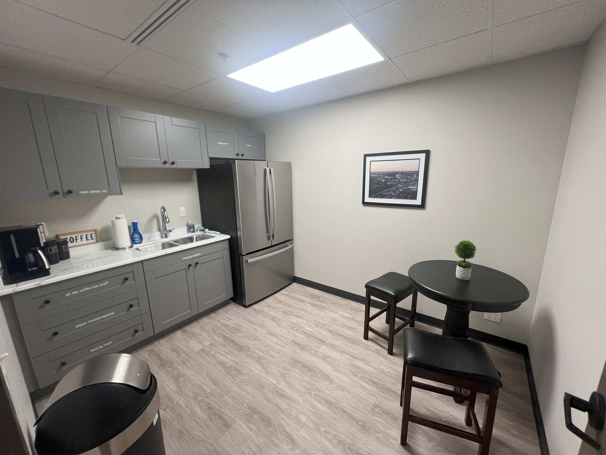 A modern small kitchen with gray cabinets, a stainless steel refrigerator, a coffee machine, a small table with two chairs, a small plant, and a framed picture on the wall.